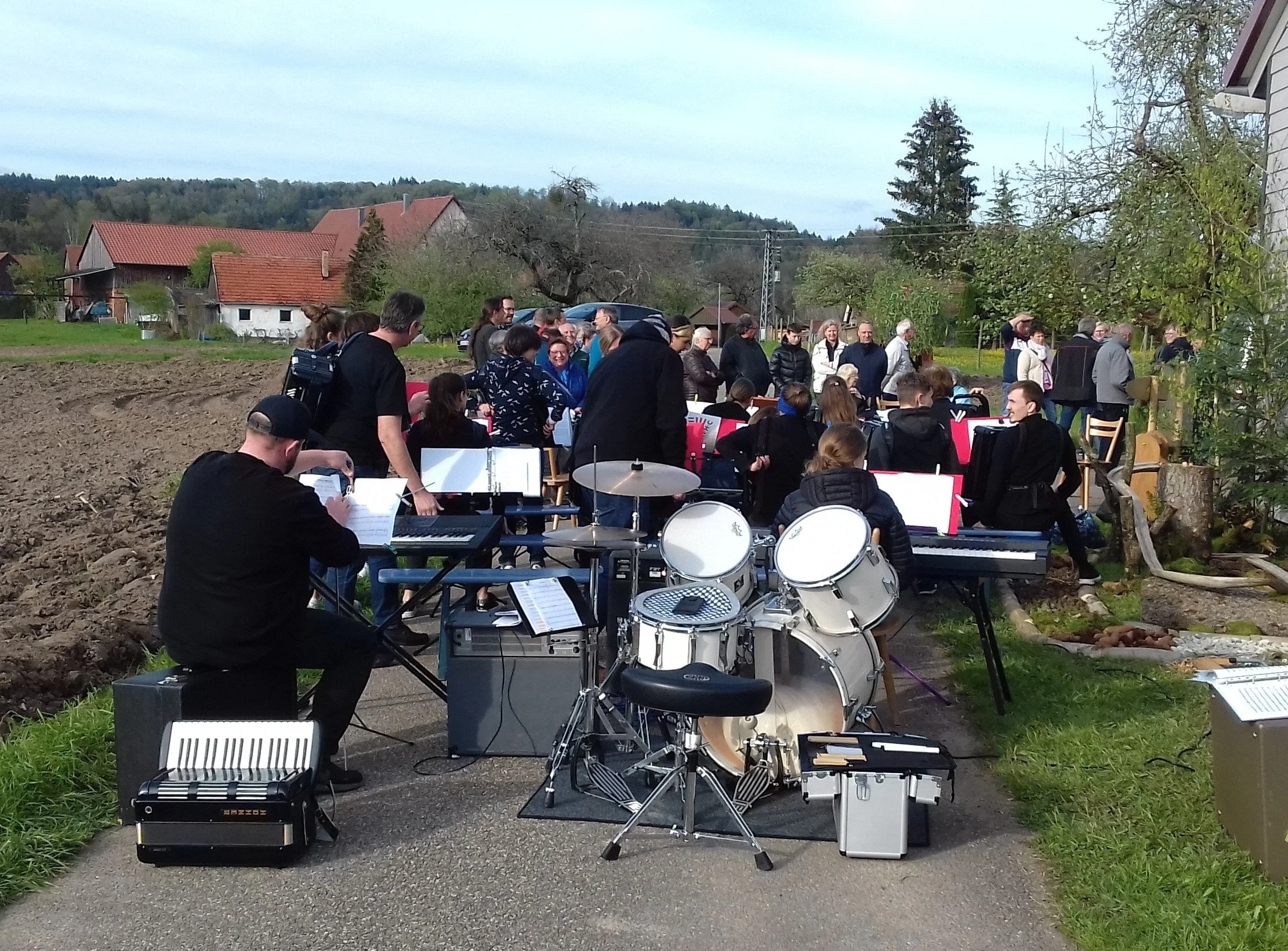 Eine Gruppe von Menschen spielt Musik im Freien, darunter ein Musiker, der an einem Keyboard sitzt, und eine Band mit Schlagzeug. Die Szenerie ist ländlich mit Häusern und Bäumen im Hintergrund.