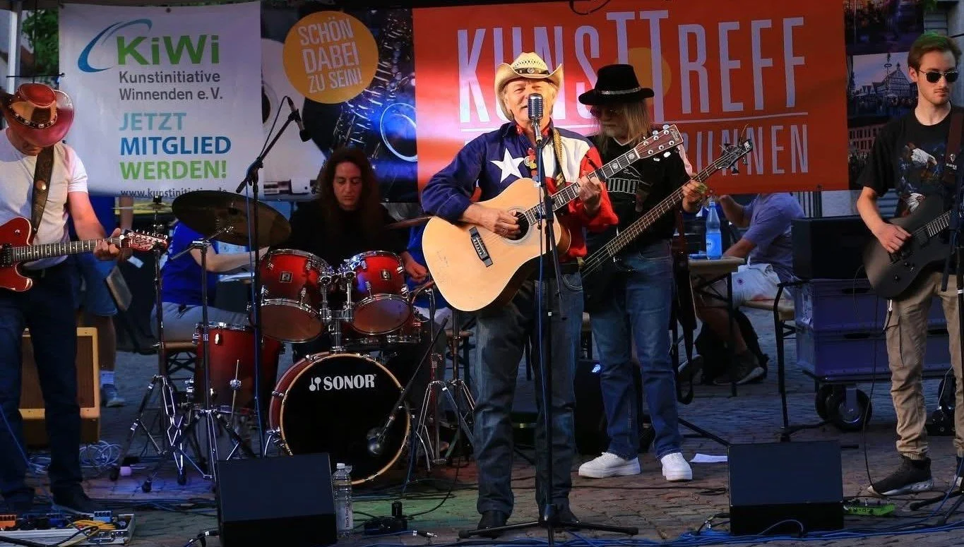 Eine Gruppe von Musikern spielt auf einer Bühne bei einem Festival. Es gibt einen Mann mit Cowboy-Hut und Gitarre, eine Drummerin, einen Mann mit E-Gitarre und einen Mann mit Bassgitarre, und ein Banner im Hintergrund.