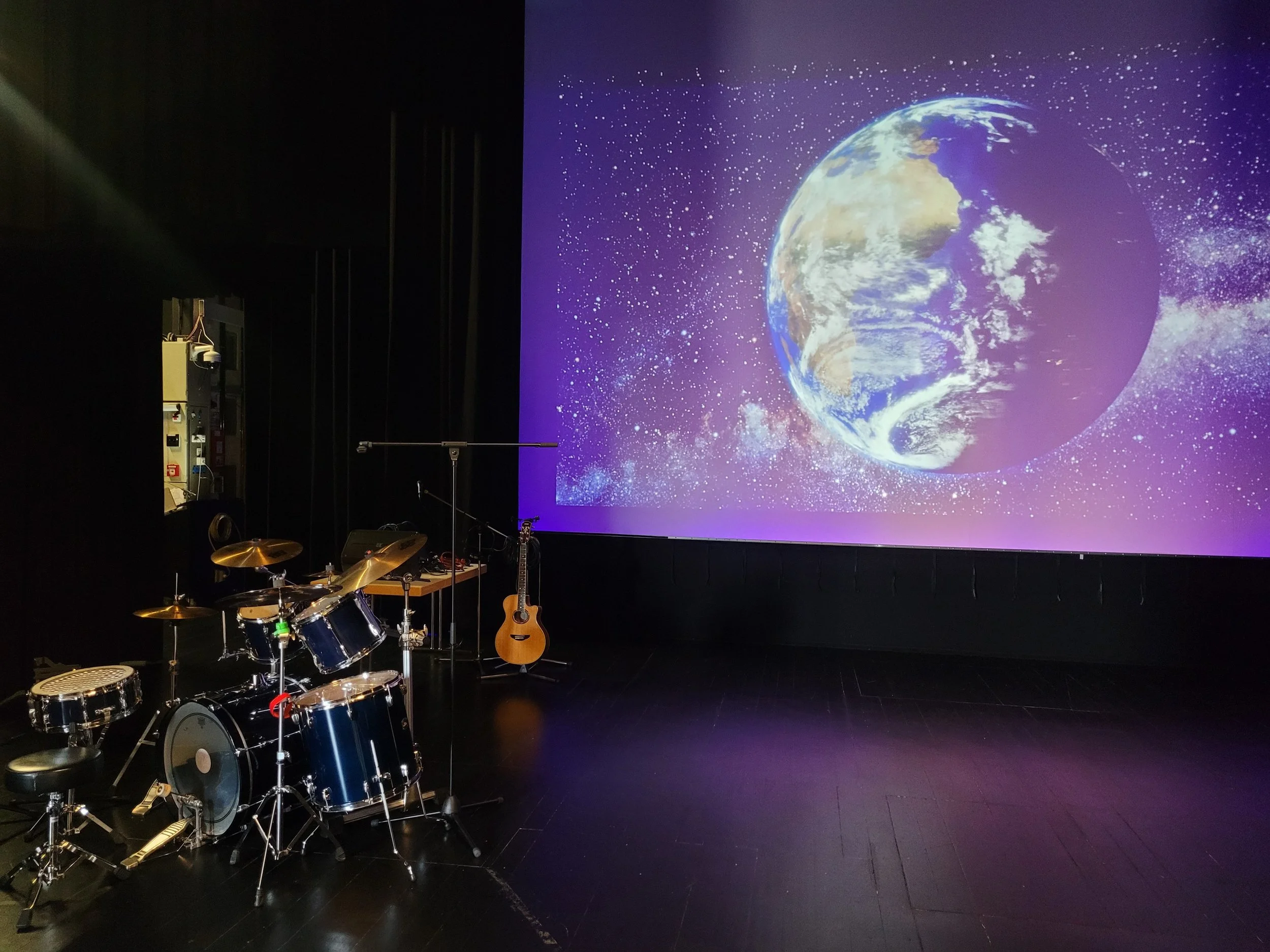 Ein leeres schwarzes Theater mit einem großen Bildschirm, auf dem die Erde im Weltall sichtbar ist. Im Vordergrund befinden sich Musikinstrumente, einschließlich eines Drumsets, einer Akustikgitarre und anderen Musikequipment.