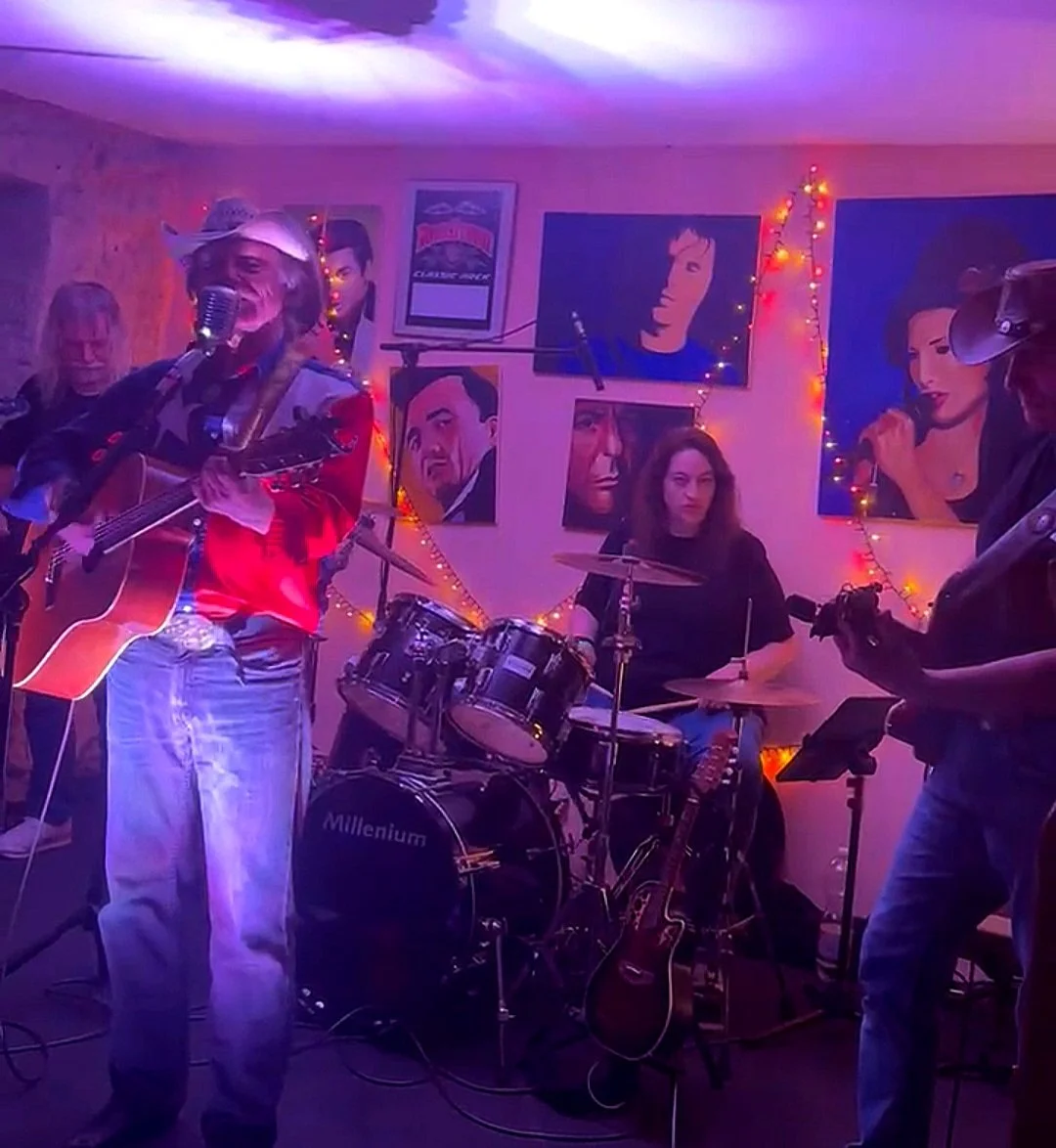 Musiker, die in einer gemütlichen, mit Pop-Art- und Filmplakaten dekorierten Bar spielen, beleuchtet von bunten Lichterketten.
