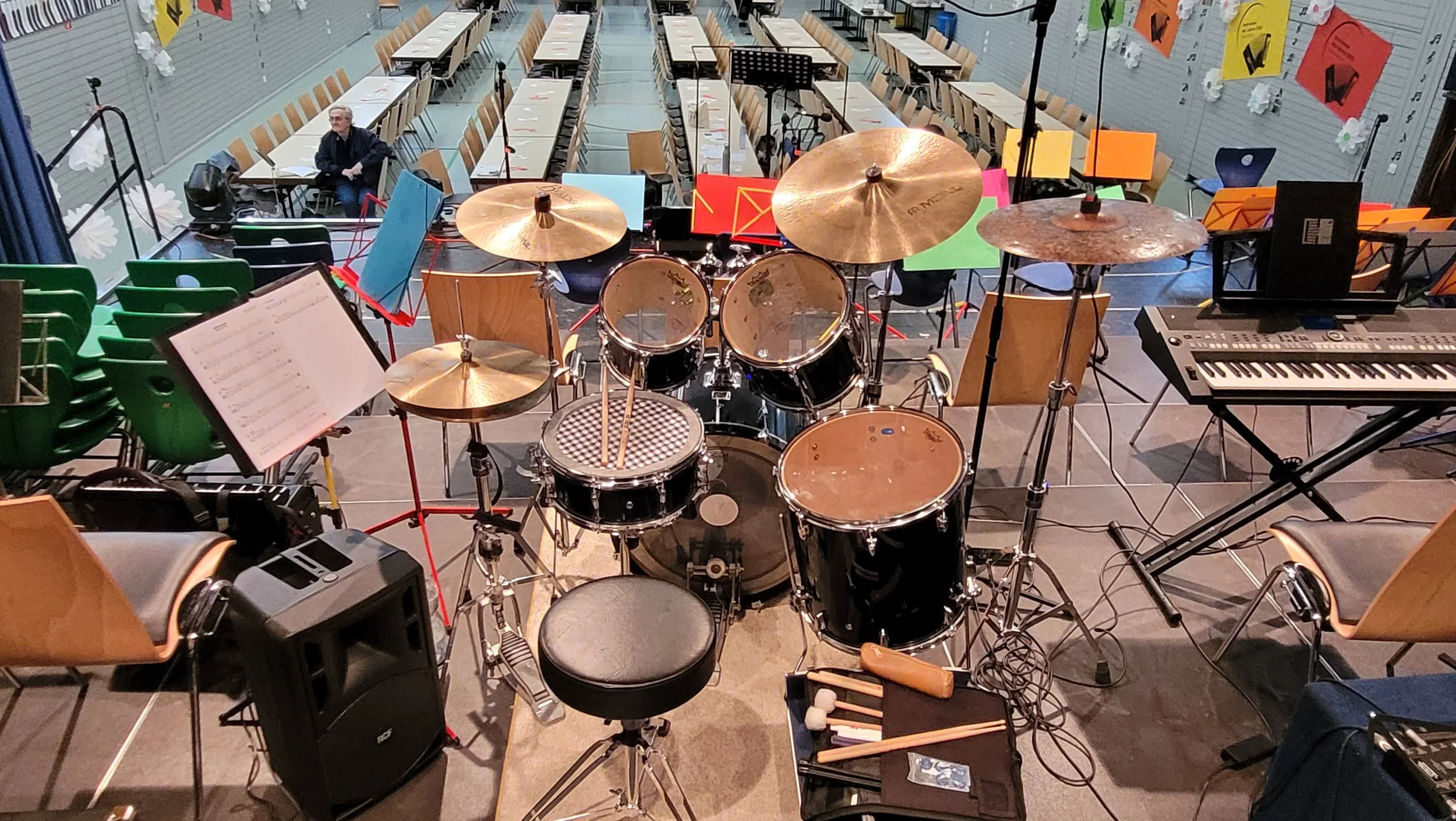 Aufnahme von einer leeren Konzertbühne mit Musikinstrumenten wie Schlagzeug, Keyboard und Notenständern, sowie Stühlen und Sitzplätzen im Hintergrund.