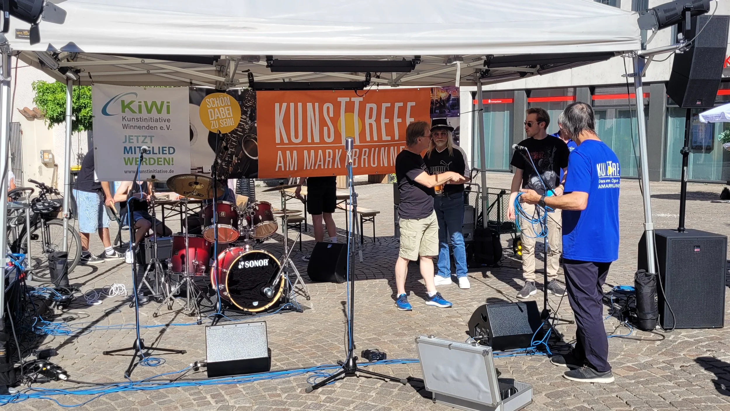 Menschen auf einer Bühne beim Musikfestival, mit Banner, Musikinstrumenten und Verstärkern im Freien.