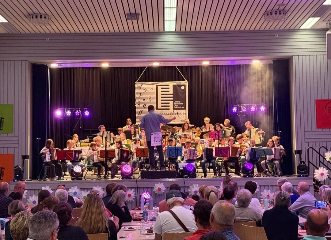 Orchester auf einer Bühne während eines Konzerts, geleitet von einem Dirigenten, mit Publikum im Saal