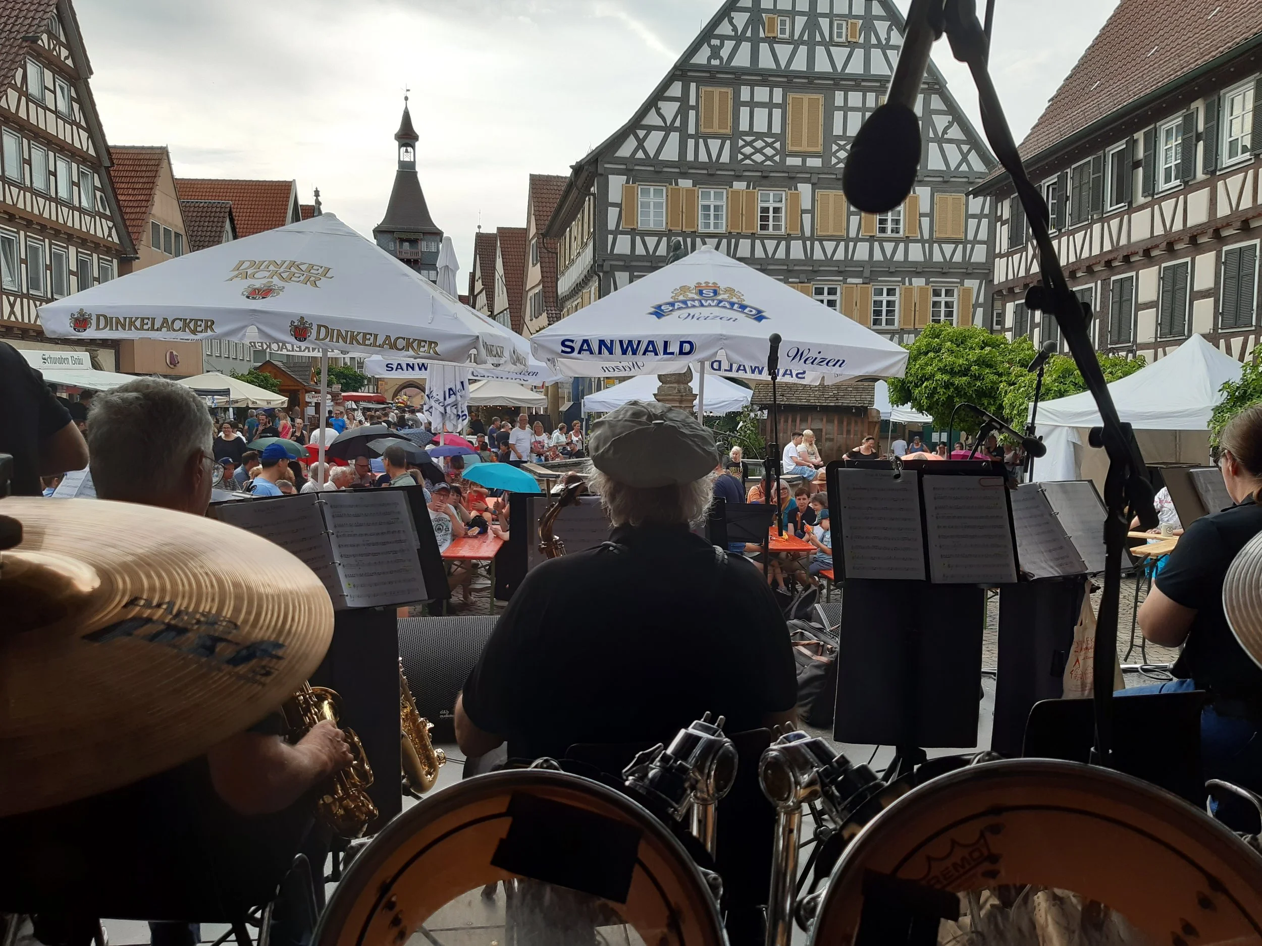 Blick auf ein Volksfest in einer historischen Stadt mit Fachwerkhäusern, klassischen Markisen, Menschen im Festzelt, Musiker im Vordergrund mit Blasinstrumenten, davor eine große Trommel. Es regnet leicht, viele Menschen haben Regenschirme, im Hintergrund ein Kirchturm und bewölkter Himmel.
