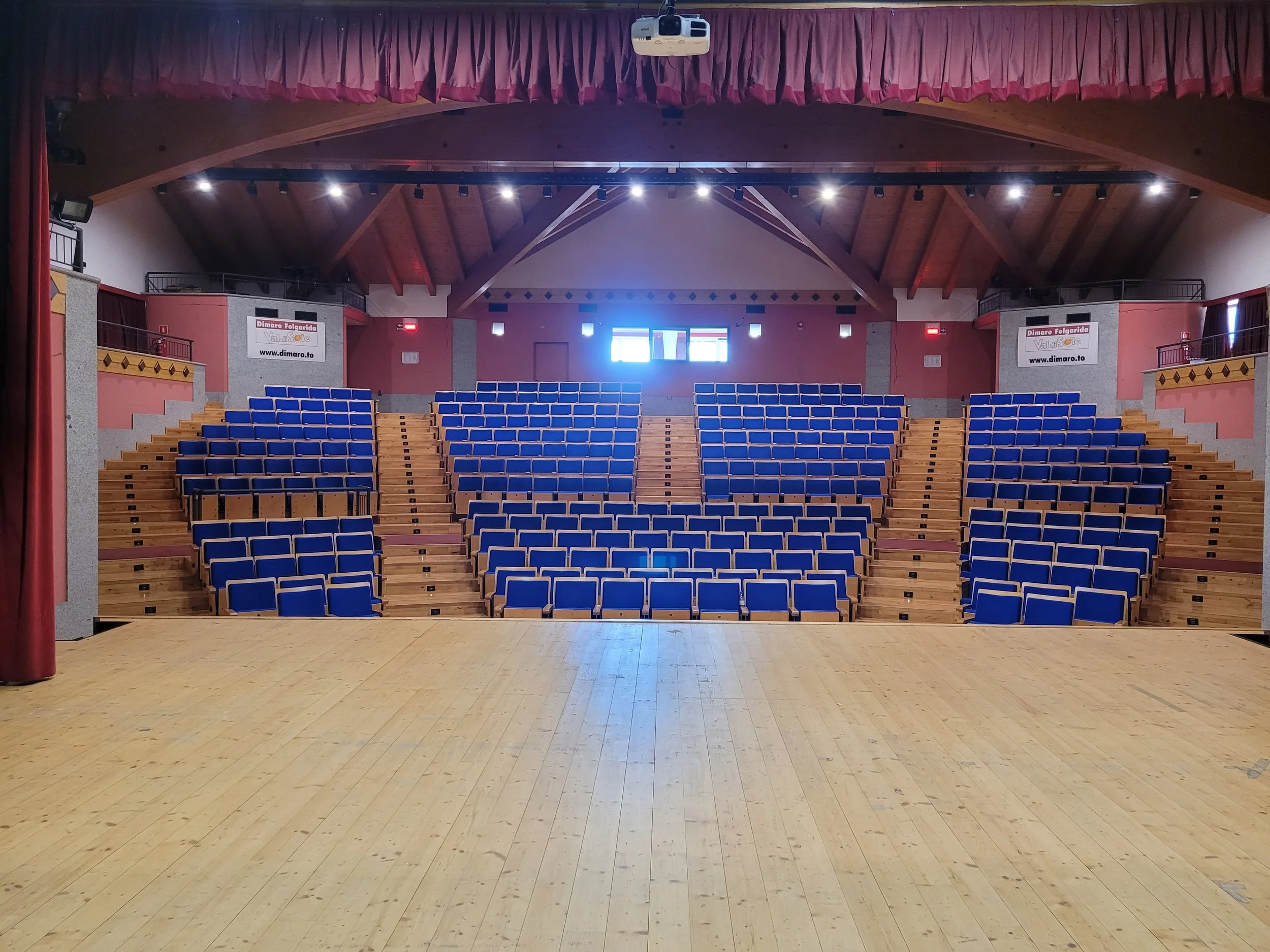 Ein leeres Auditorium mit blauen Sitzreihen, einem Holzfußboden und einem hohen, gewölbten Holzdecke. Am hinteren Ende des Raumes sind Fenster, die Licht hereinlassen, und es hängen Scheinwerfer zur Beleuchtung.