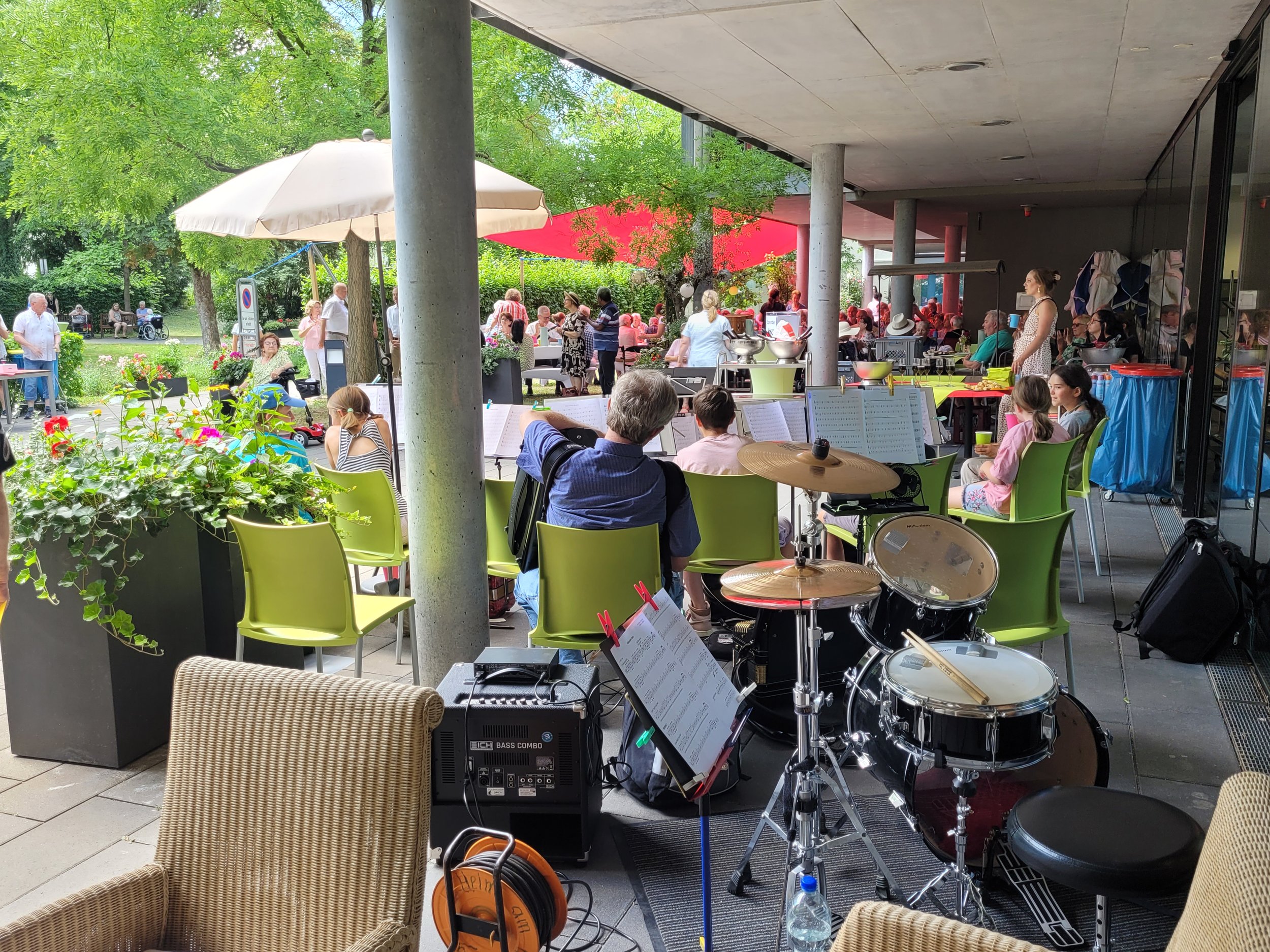 Ein Veranstaltungsort im Freien mit vielen Menschen, die an einem bunten, sonnigen Tag zusammensitzen. Es gibt einen Musikbereich mit Schlagzeug und Musikanlage im Vordergrund.