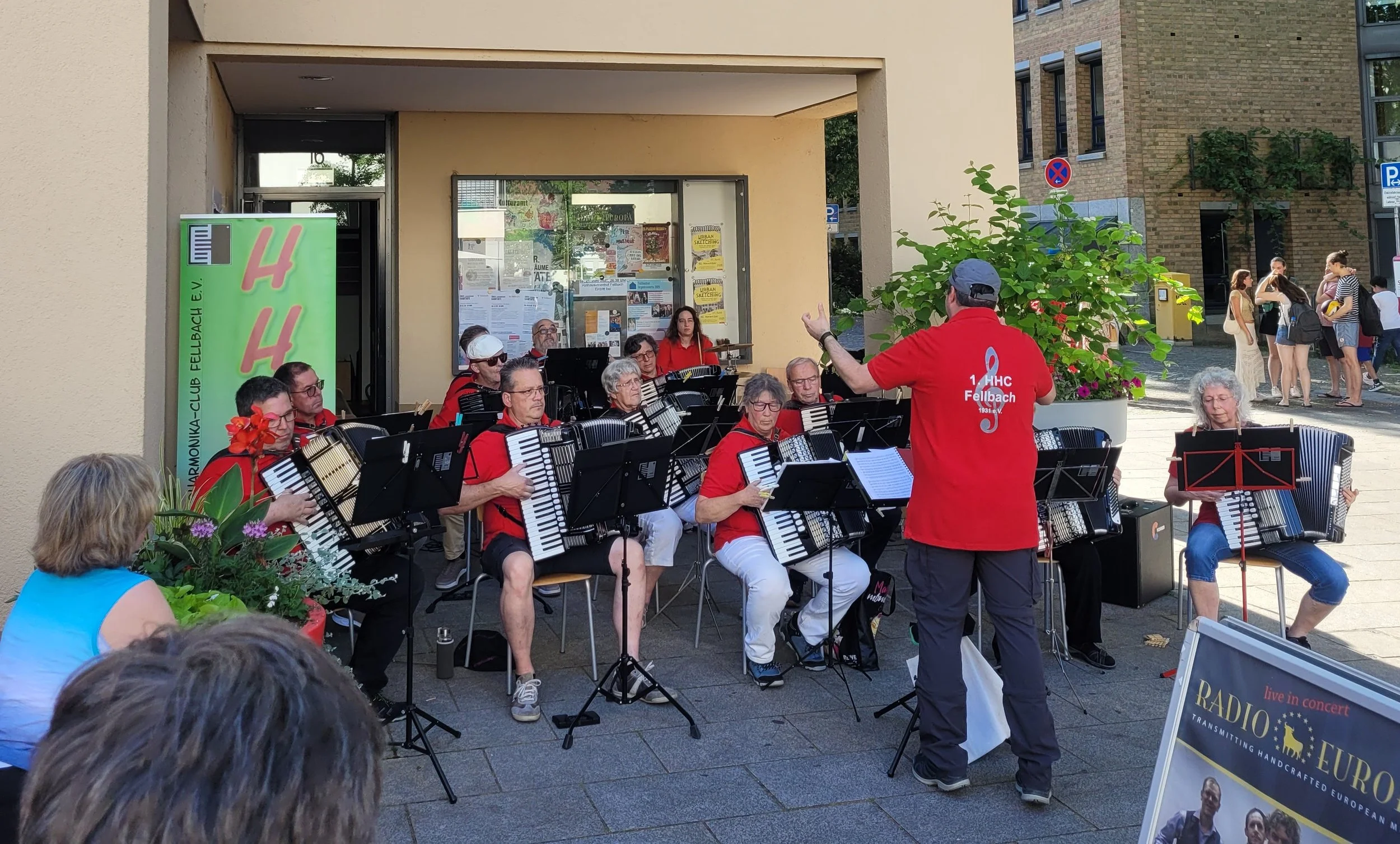 Eine Gruppe von Musikern spielt auf einer Bühne im Freien. Es handelt sich um ein Orchester, das auf einem Gehweg vor einem Gebäude auftritt. Ein Dirigent führt die Musiker und im Hintergrund sind Zuschauer zu sehen. Die Musiker tragen rote Hemden und es gibt einen Stand mit Plakaten und Blumen.