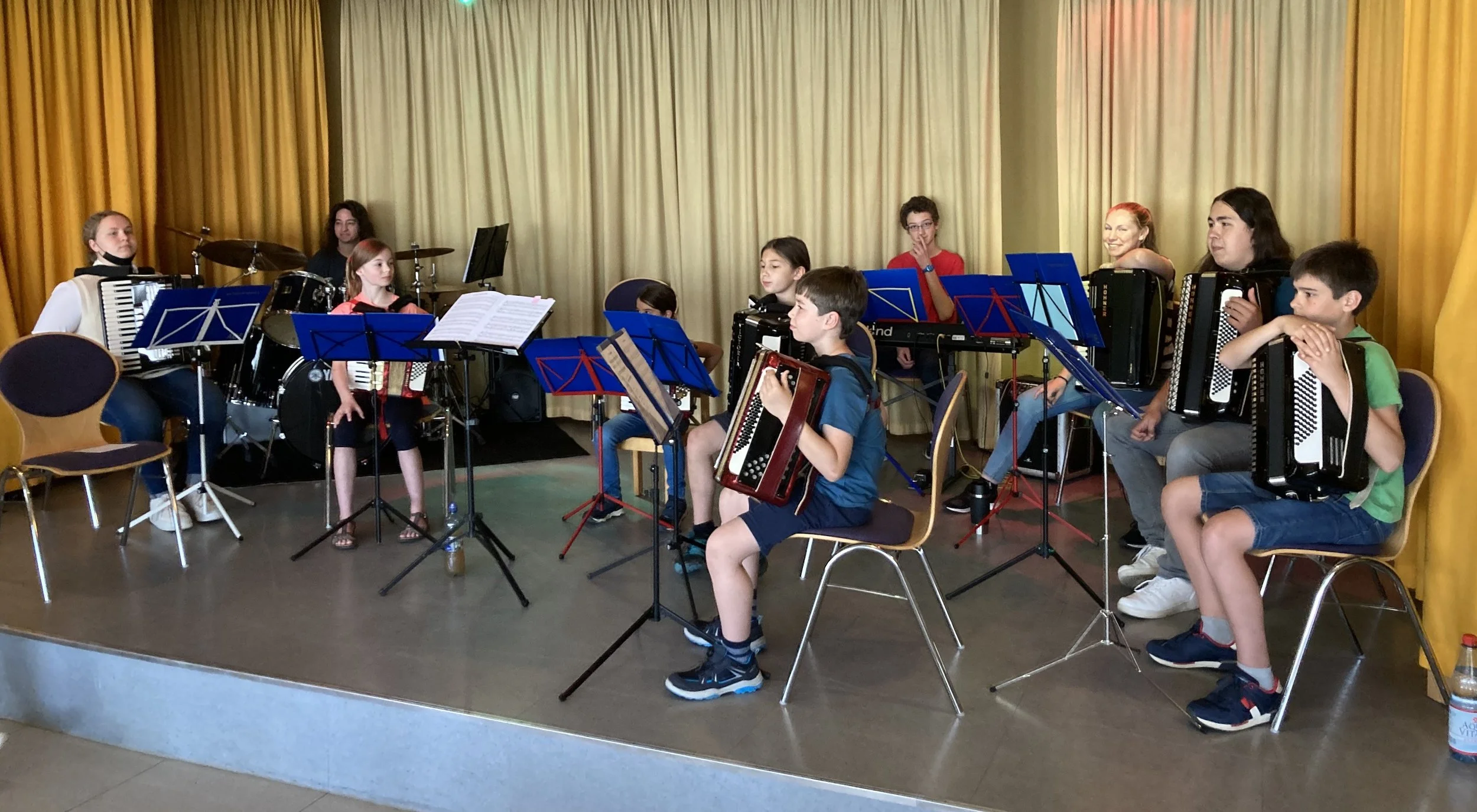 Jungen und Mädchen auf einer Bühne, spielen Akkordeon, mit einem Schlagzeug im Hintergrund und einem Notenständer in der Mitte, vor goldgelben Vorhängen.