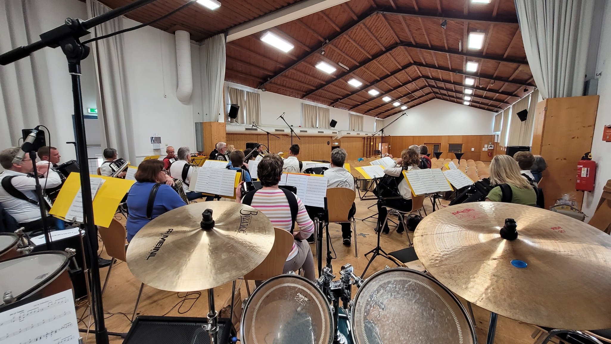 Orchesterprobe in einem großen Saal, aufgenommen vom Schlagzeug aus. Musiker sitzen mit ihren Instrumenten und Notenblättern, die Bühne ist vorbereitet, mit Mikrofonen und weiteren technischen Geräten. Es herrscht eine ruhige, konzentrierte Atmosphäre.