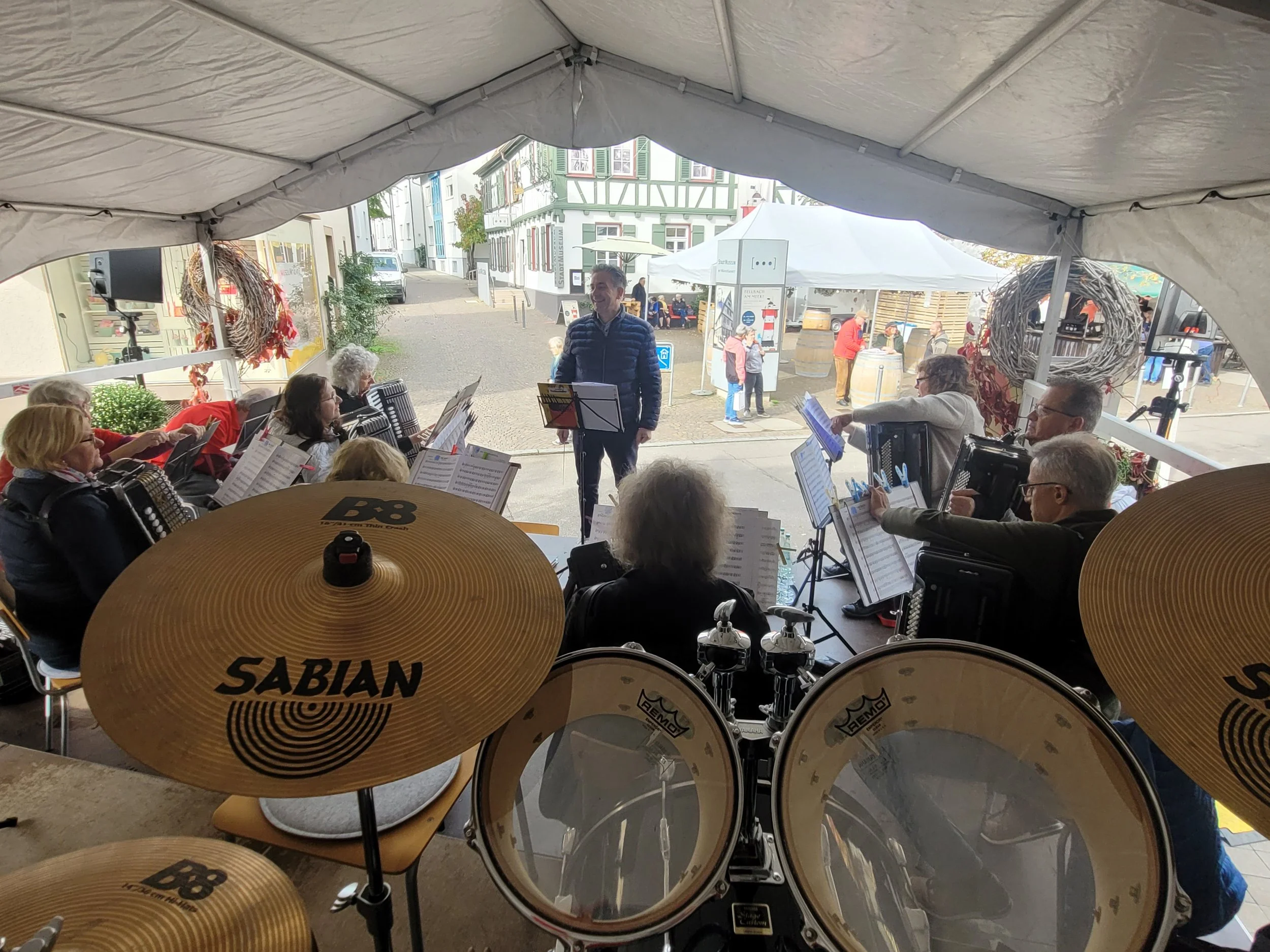 Ein Blick aus einem Zelt auf eine Straße mit Menschen, die sich versammeln, einige sitzen auf Bänken, andere gehen herum. In dem Zelt sind Musikinstrumente, darunter Schlagzeuge und eine Gitarre, sichtbar.
