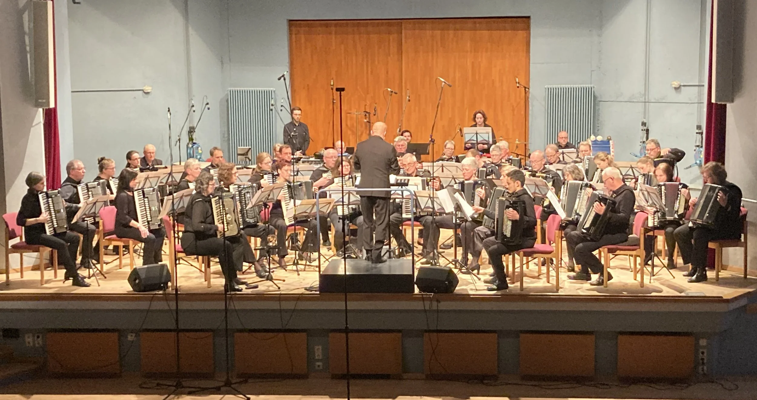 Ein Orchester mit Musikern, die Akkordeon spielen, wird von einem Dirigenten geleitet. Das Konzert findet in einem Raum mit Holz-Falttür und Mikrofonständern statt.
