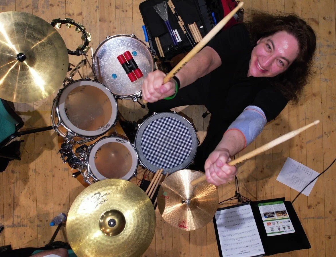 Eine Person sitzt hinter einem Drum-Set, das von oben aufgenommen wurde, und schwingt mit Schlagstöcken in den Händen. Das Setup umfasst Trommeln und Becken, sowie eine Notenmappe auf einer Musikstativ. Der Boden ist aus Holz.