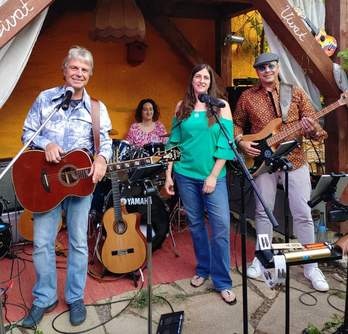 Musiker mit Gitarren und Schlagzeug auf einer Bühne, zwei Männer und eine Frau singen und spielen, im Hintergrund ein Schlagzeug, ein Musikständer, eine Außenkulisse mit Holzrahmen und gelber Wand.