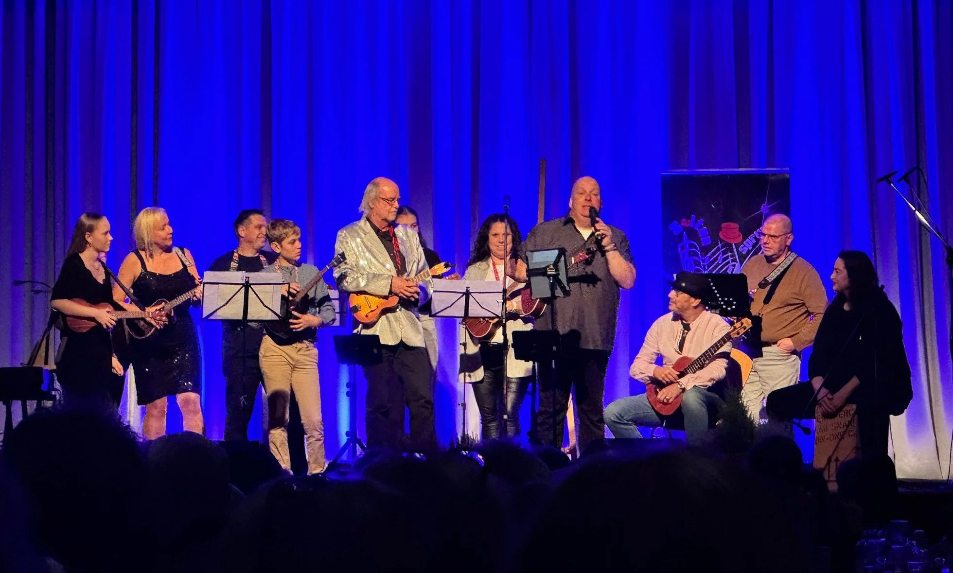 Eine Gruppe von Musikern und Sängerinnen auf einer Bühne mit blauen Vorhängen im Hintergrund; einige spielen Gitarre, andere singen oder halten Instrumente.