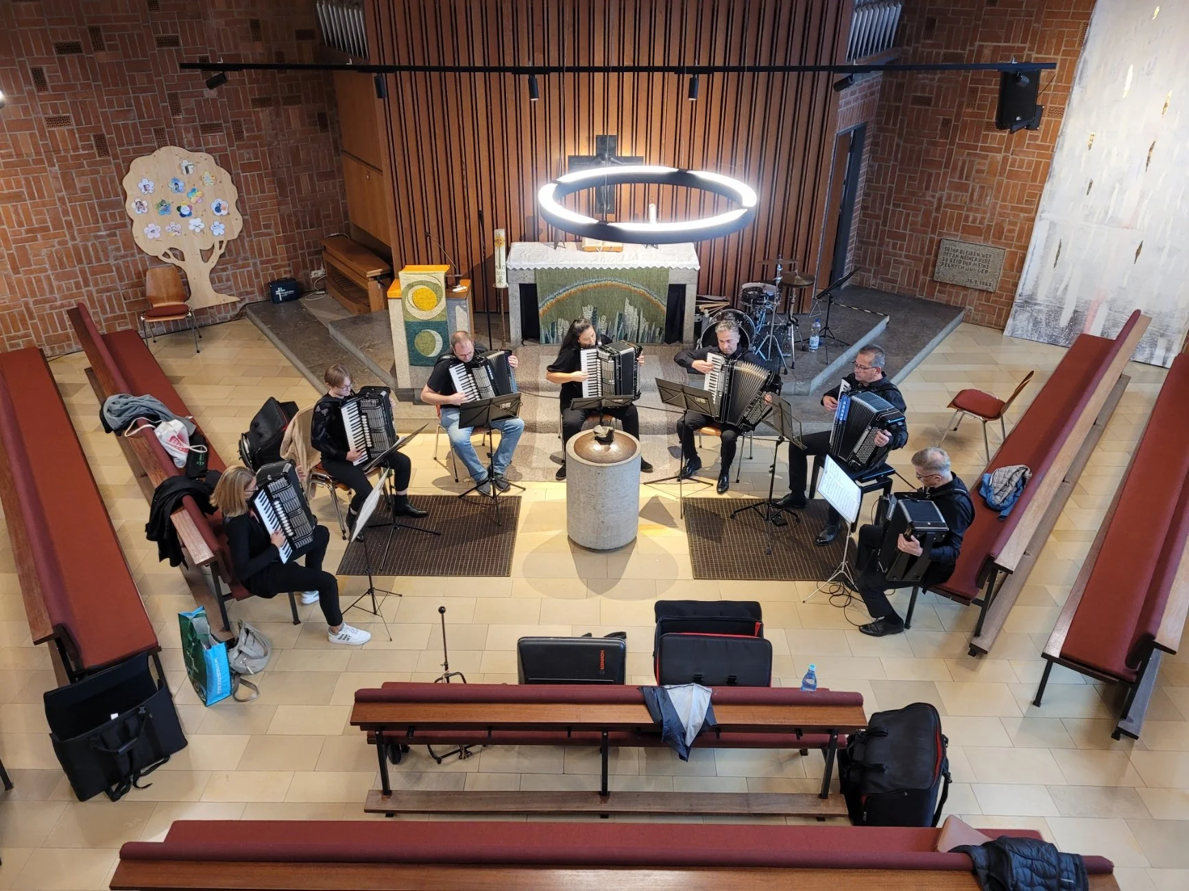 Ein Chor von sieben Personen, die Akkordeon spielen, befindet sich auf einer kleinen Bühne in einer Kirche. Die Bühne hat einen Altar im Hintergrund und ist mit Notenständern ausgestattet. Es gibt einen Kreis aus moderner Beleuchtung über der Bühne. Die Kirche hat Ziegelwände und Holz-Bänke, die nach oben ansteigen.