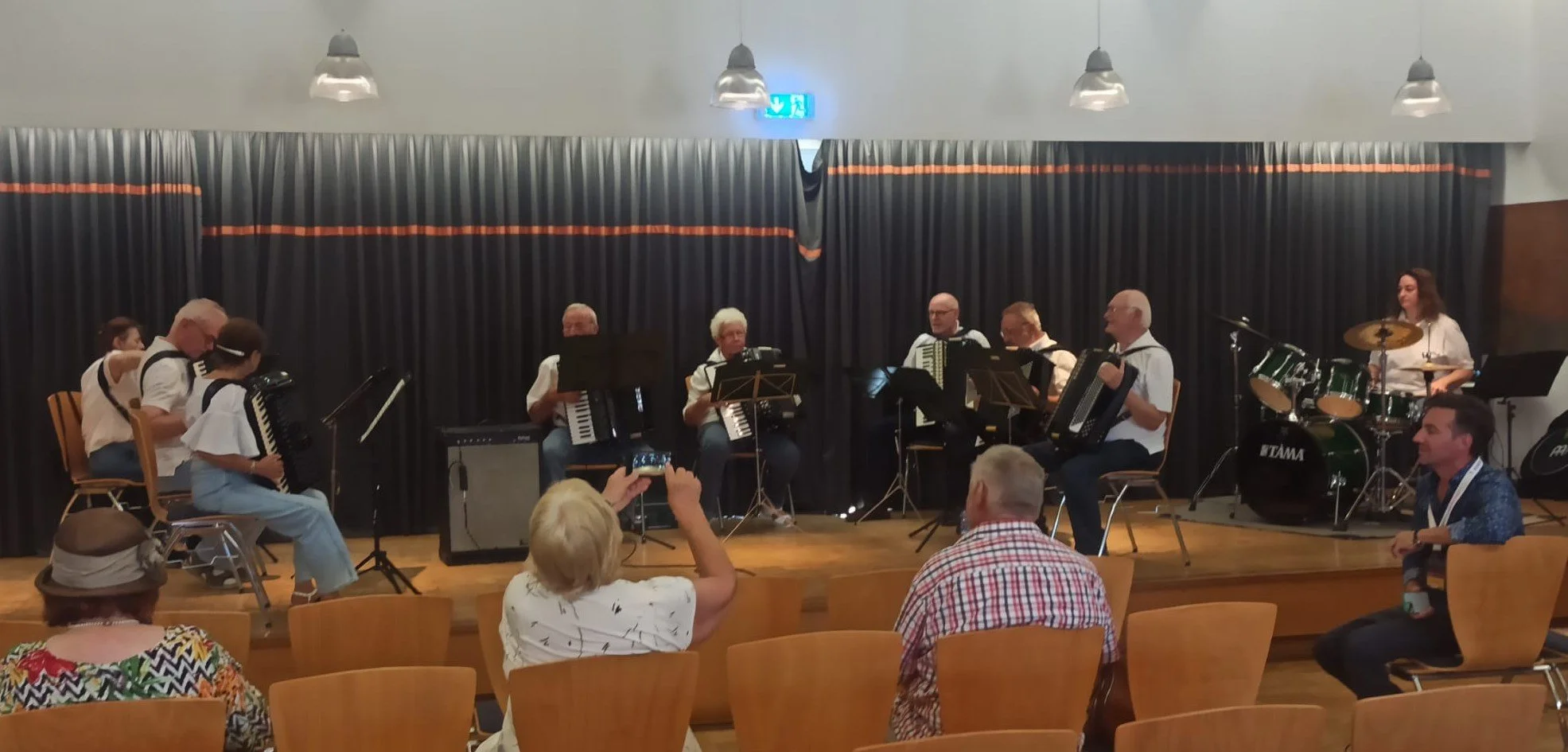 Musiker spielen auf einer Bühne, begleitet von einer Schlagzeugerin, während das Publikum zuschaut. Es gibt verschiedene Instrumente wie Akkordeons, Schlagzeug und E-Gitarre.