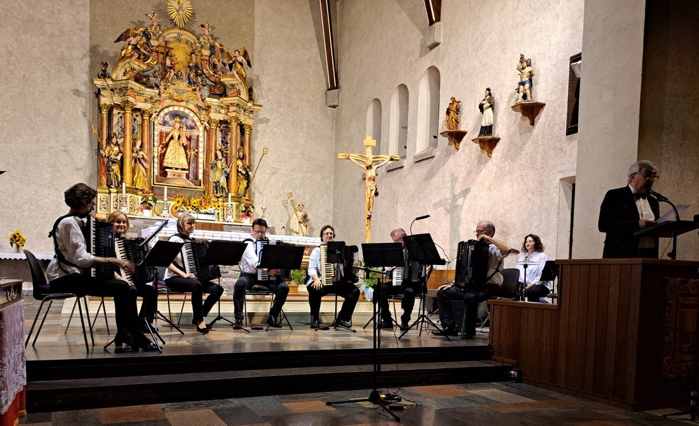 Eine Gruppe von Musikern spielt auf einer Bühne in einer Kirche, im Hintergrund ein Altar mit goldverzierten Figuren, eine goldene Christusfigur am Kreuz und einige Heiligenfiguren auf kleinen Wandregalen.