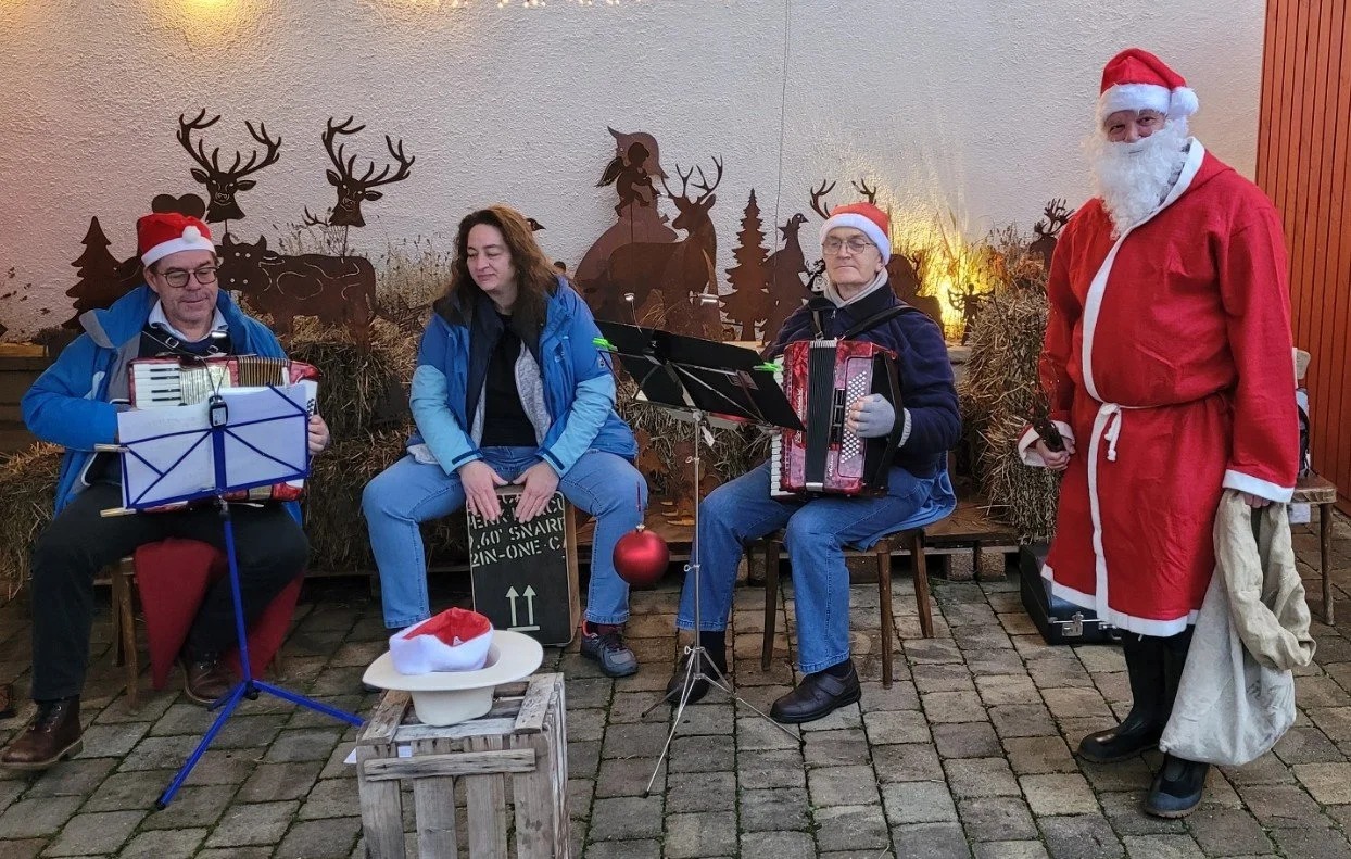 Vier Personen in Weihnachtskostümen, zwei mit Weihnachtsmützen, sitzen oder stehen vor einer weihnachtlichen Deko mit Rentiere und Tannenbäumen, einige spielen Musikinstrumente und eine Person trägt einen roten Santa-Kostüm.