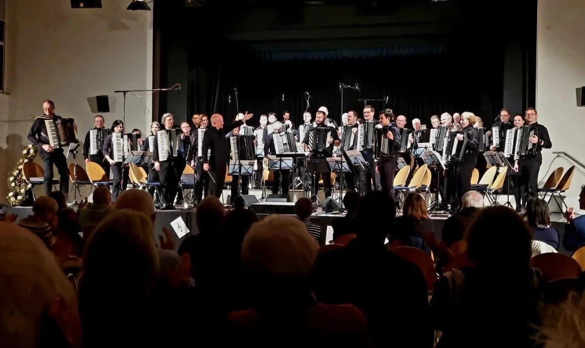 Ein Orchester auf einer Bühne, das von einem Dirigenten geleitet wird. Das Orchester besteht aus Musikern, die Akkordeons spielen. Das Publikum sitzt im Zuschauerraum und schaut zu.