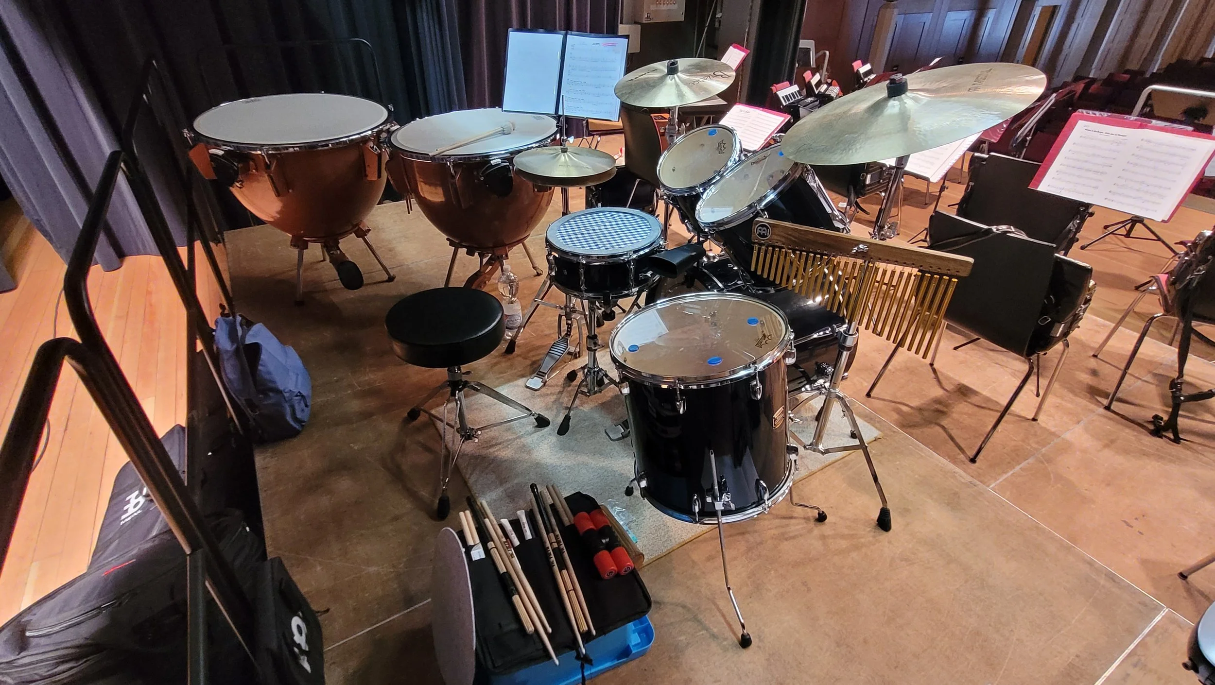 Ein Schlagzeug-Set auf einer Bühne mit Notenständern und Musiknoten. Es gibt Trommeln, Becken und verschiedene Percussion-Instrumente.