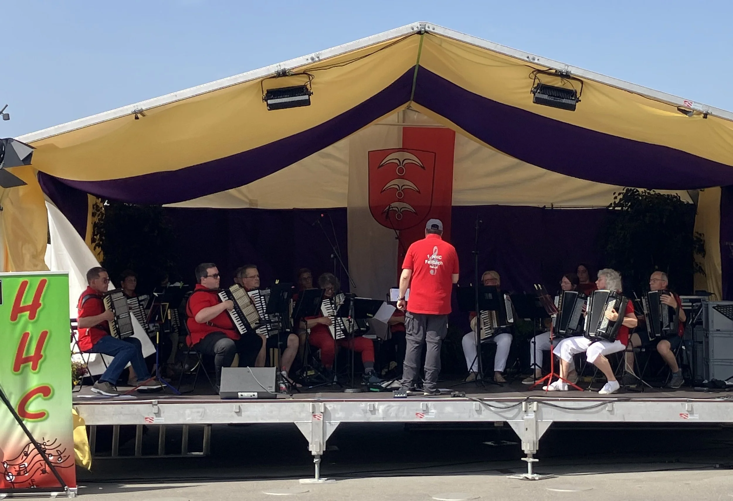 Ein Orchester spielt auf einer Bühne im Freien während eines Festivals oder einer Veranstaltung, unter einem großen gelben und lila Zelt. Es ist Tageszeit, und die Musiker tragen rote Hemden, einige von ihnen spielen Akkordeons, während ein Dirigent anwesend ist.