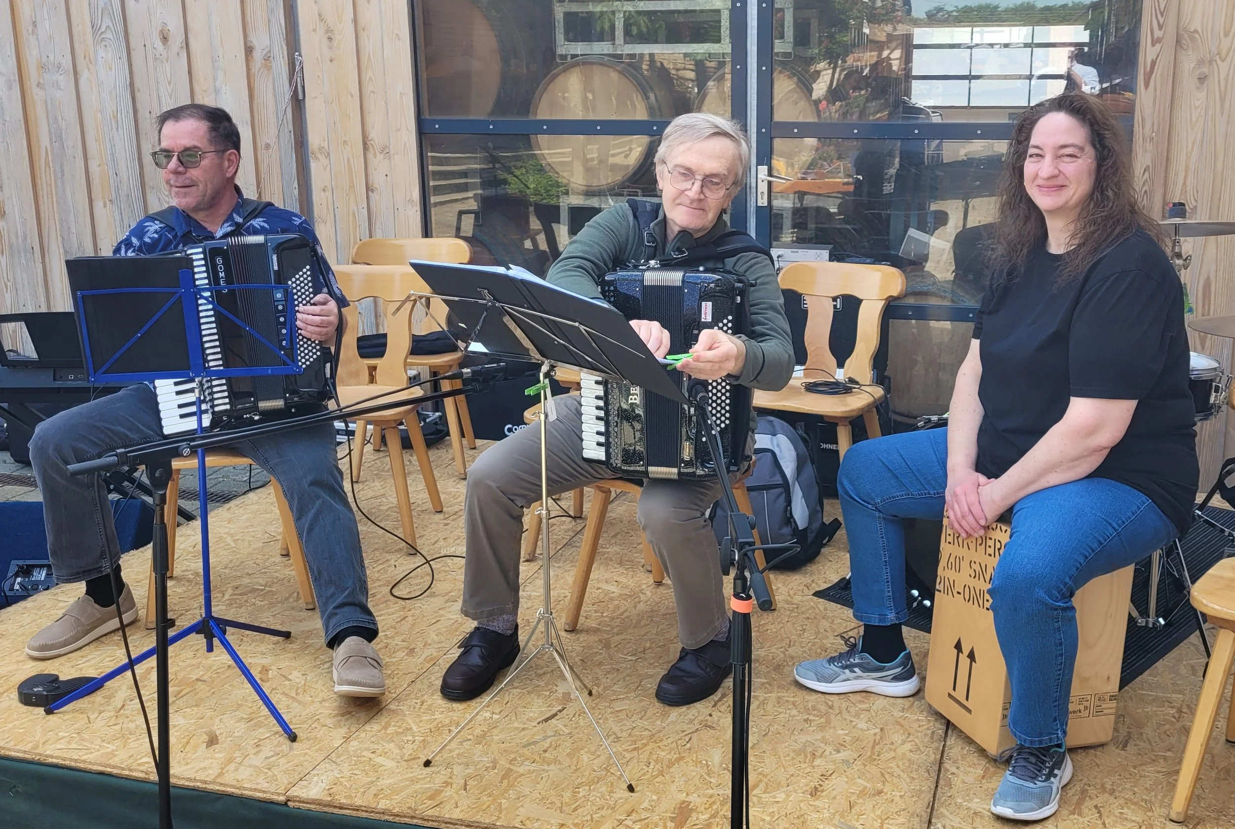 Drei Menschen sitzen auf einer Bühne, die aus Holz besteht. Zwei Männer spielen Akkordeon und eine Frau spielt Cajon, während sie Musik machen. Es gibt Notenständer und Musikinstrumente um sie herum.