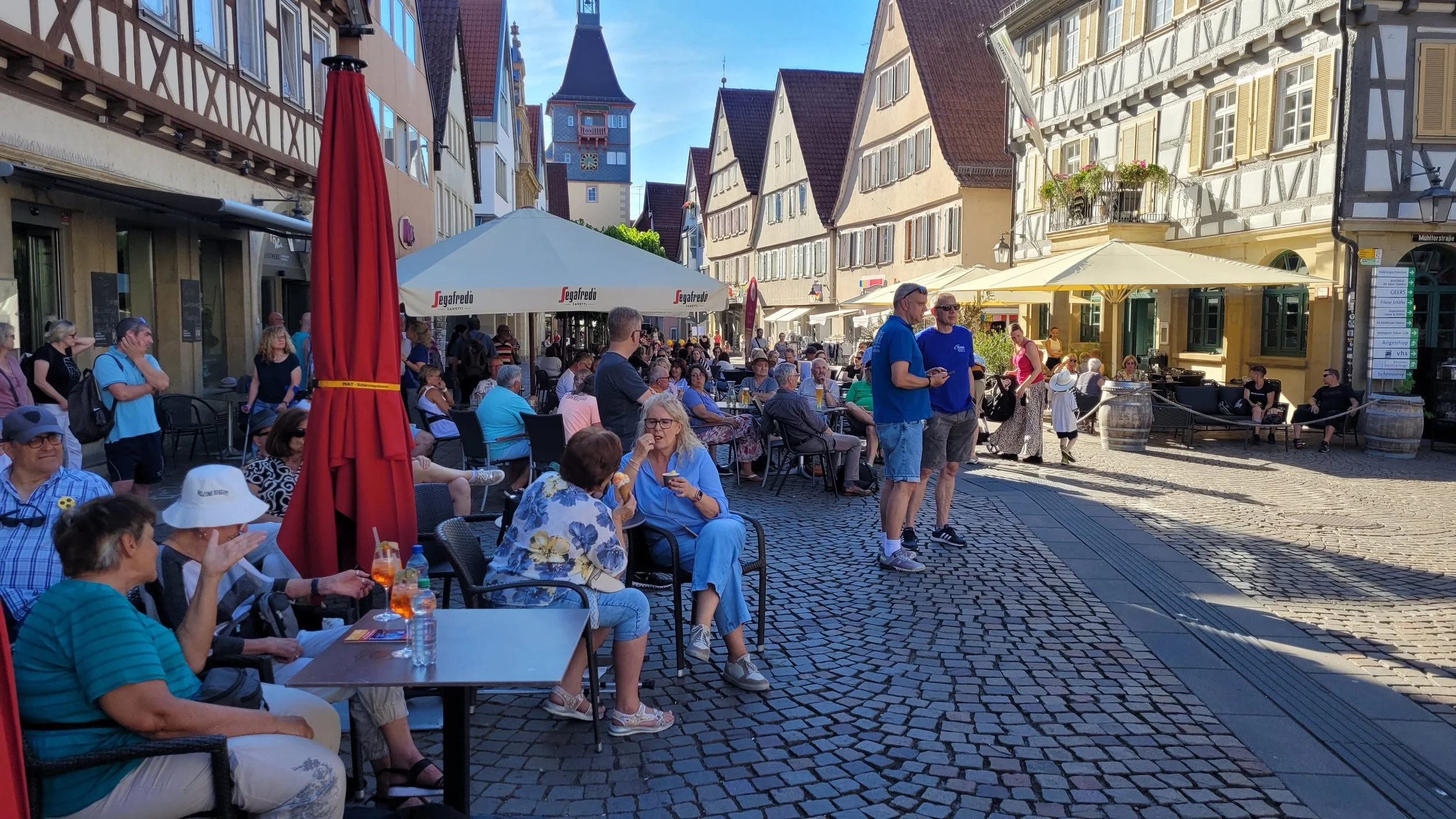 Ein belebter Stadtplatz mit vielen Menschen, die bei schönem Wetter unter Sonnenschirmen sitzen und sich unterhalten, umgeben von historischen Gebäuden mit bunten Fassaden und einem Kirchturm im Hintergrund.