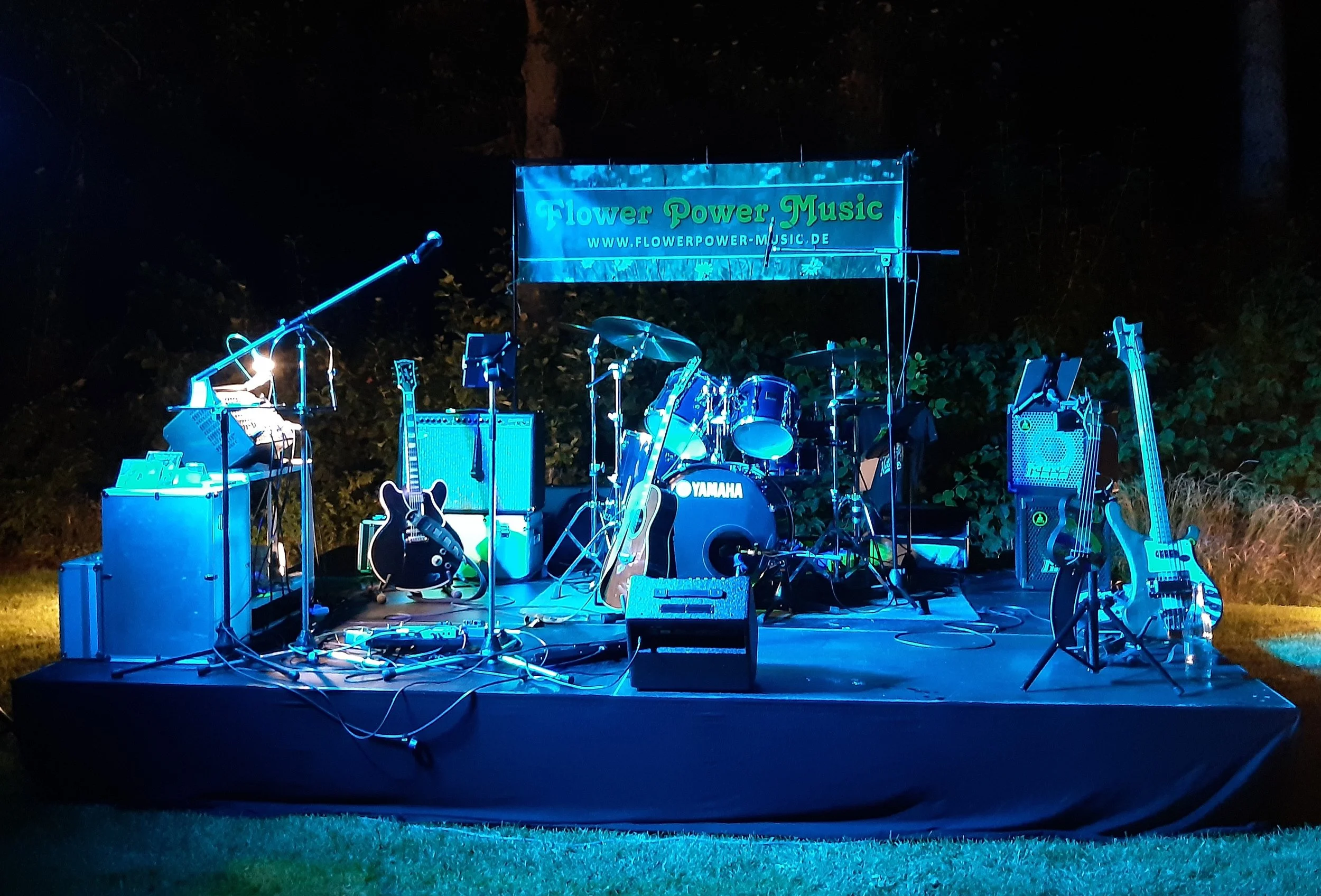 Bühne im Freien mit Musikinstrumenten wie Gitarren, Schlagzeug und Mikrofon, beleuchtet bei Nacht, mit einem Banner, das 'Flower Power Music' und eine Webseite zeigt.