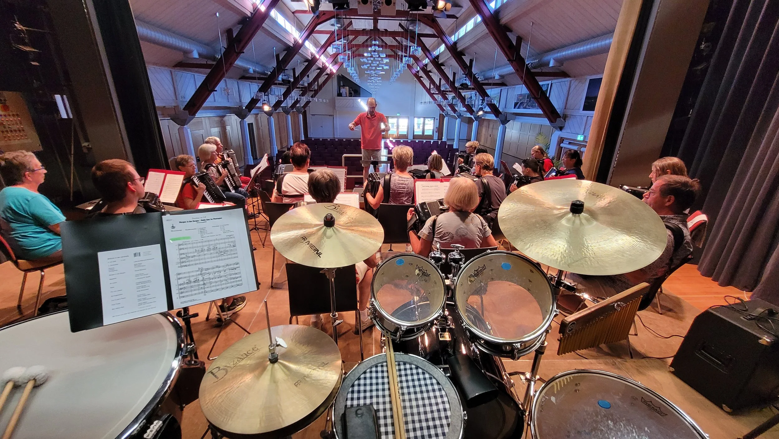 Ein Orchester spielt in einem Saal, mit einem Dirigenten an der Front, während die Musiker Instrumente spielen, darunter auch Akkordeons. Blick von hinten auf das Schlagzeug, das im Vordergrund zu sehen ist.