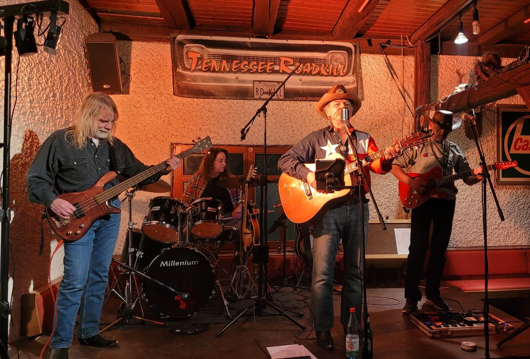 Eine Band mit fünf Musikern spielt auf einer Bühne. Der Musiker in der Mitte trägt einen Cowboyhut und spielt Gitarre, während die anderen Instrumente spielen, darunter Bass, Schlagzeug und eine zweite Gitarre.