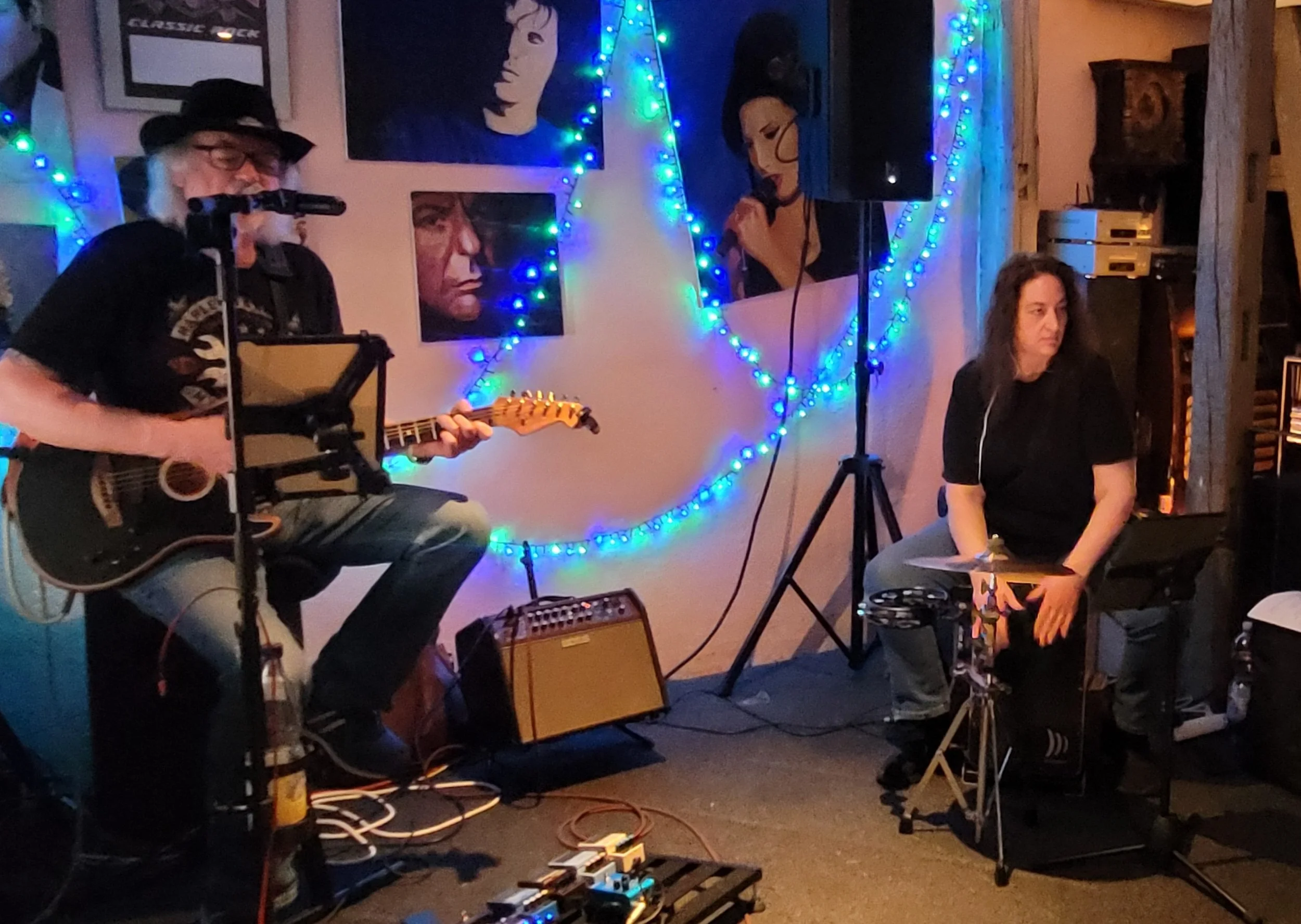 Musiker auf einer Bühne mit Gitarre und Mikrofon. Eine Personsitzt auf einer Cajon und hat ein Becken. Bunte Lichterdekorationen an der Wand,