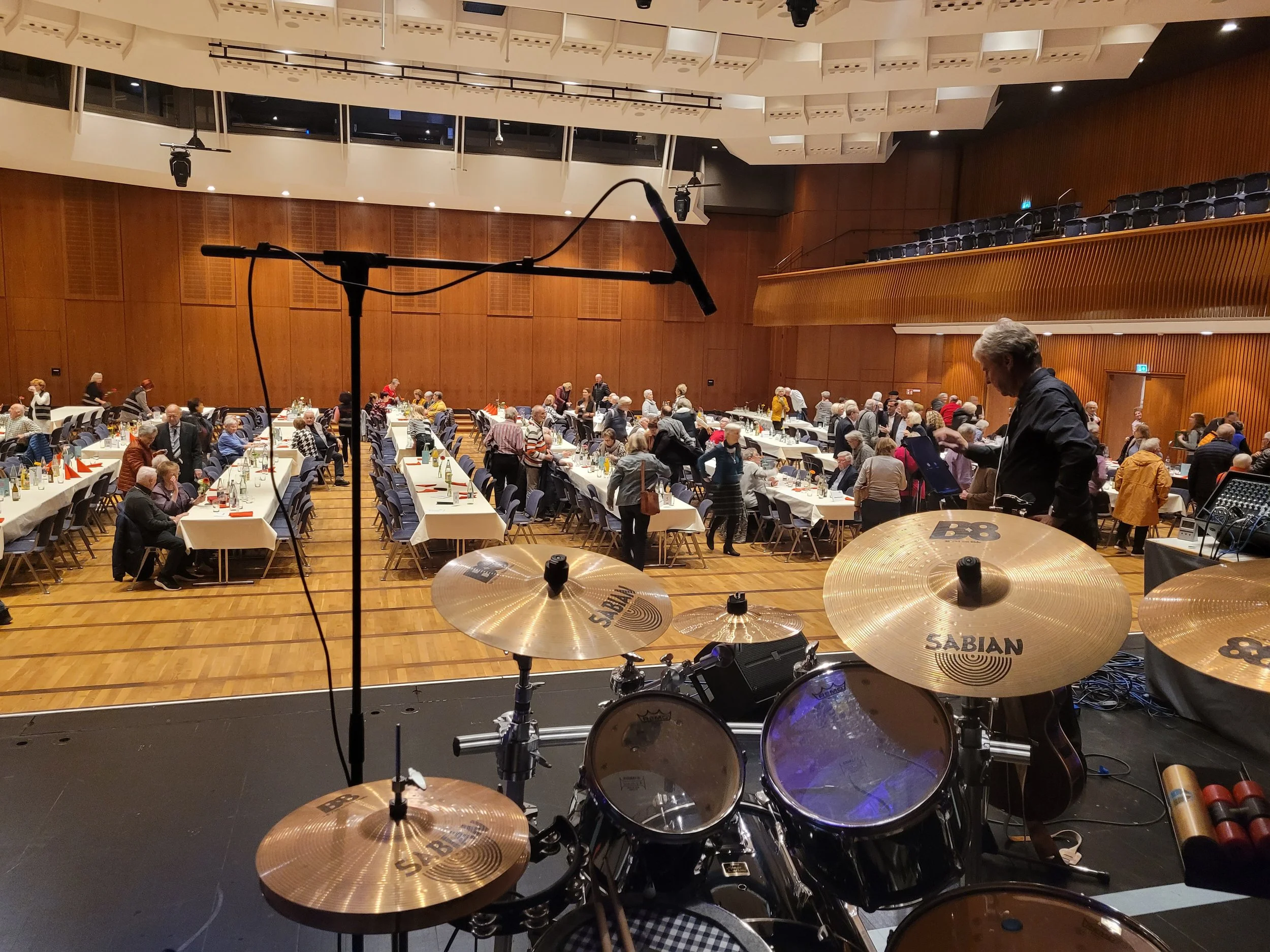 Blick von einem musikalischen Auftritt auf eine große, voll besetzte Veranstaltungshalle mit Tischen, auf denen Getränke und Servietten stehen. Im Vordergrund ist ein Schlagzeug zu sehen, und auf der Bühne sind Mikrofone angebracht.