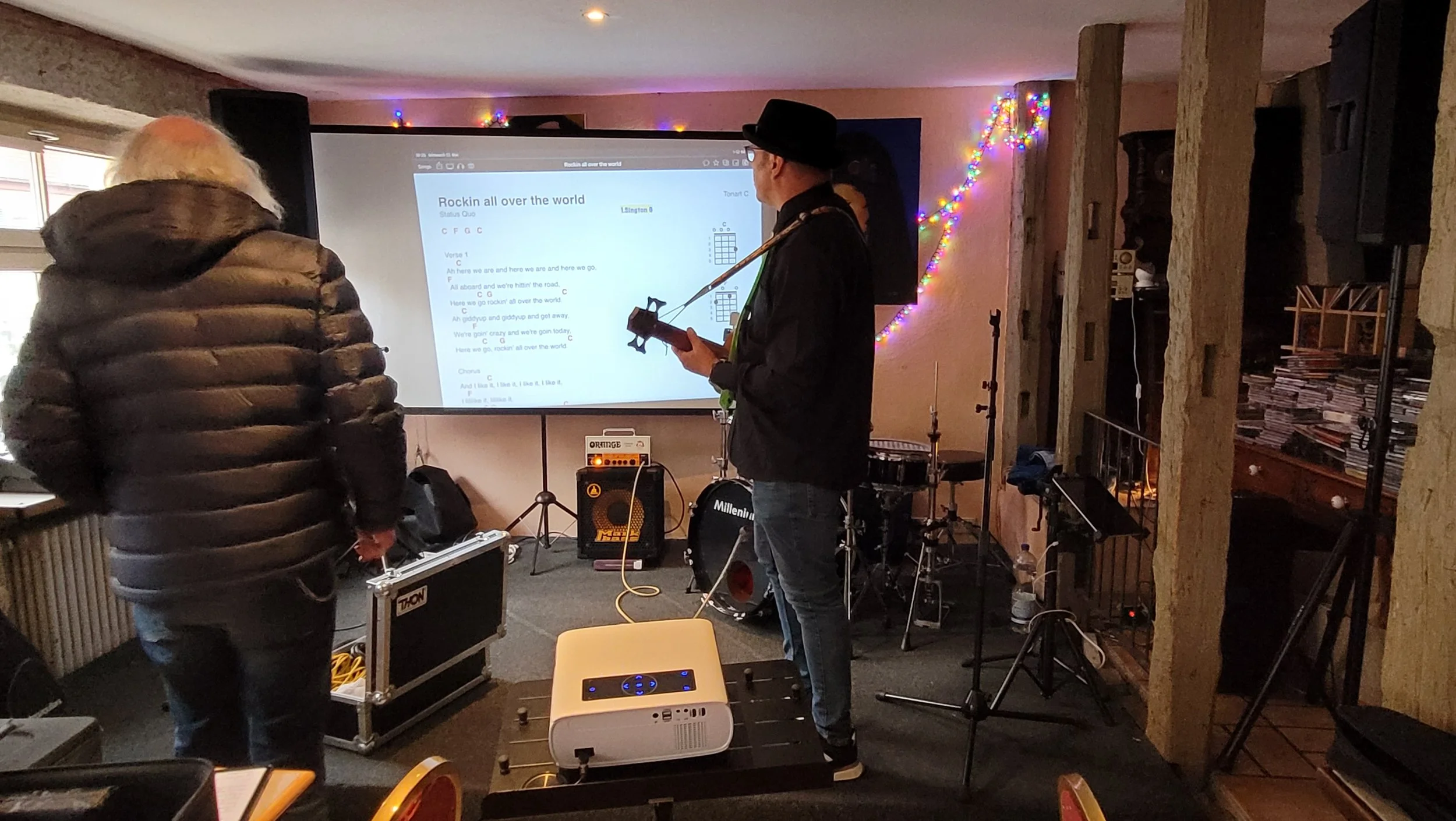 Zwei Männer beim Aufbau für ein Karaoke-Event, im Hintergrund ein Bildschirm mit Songtext, bunte Weihnachtslichter an der Wand und einige Musikinstrumente und Equipment im Raum.