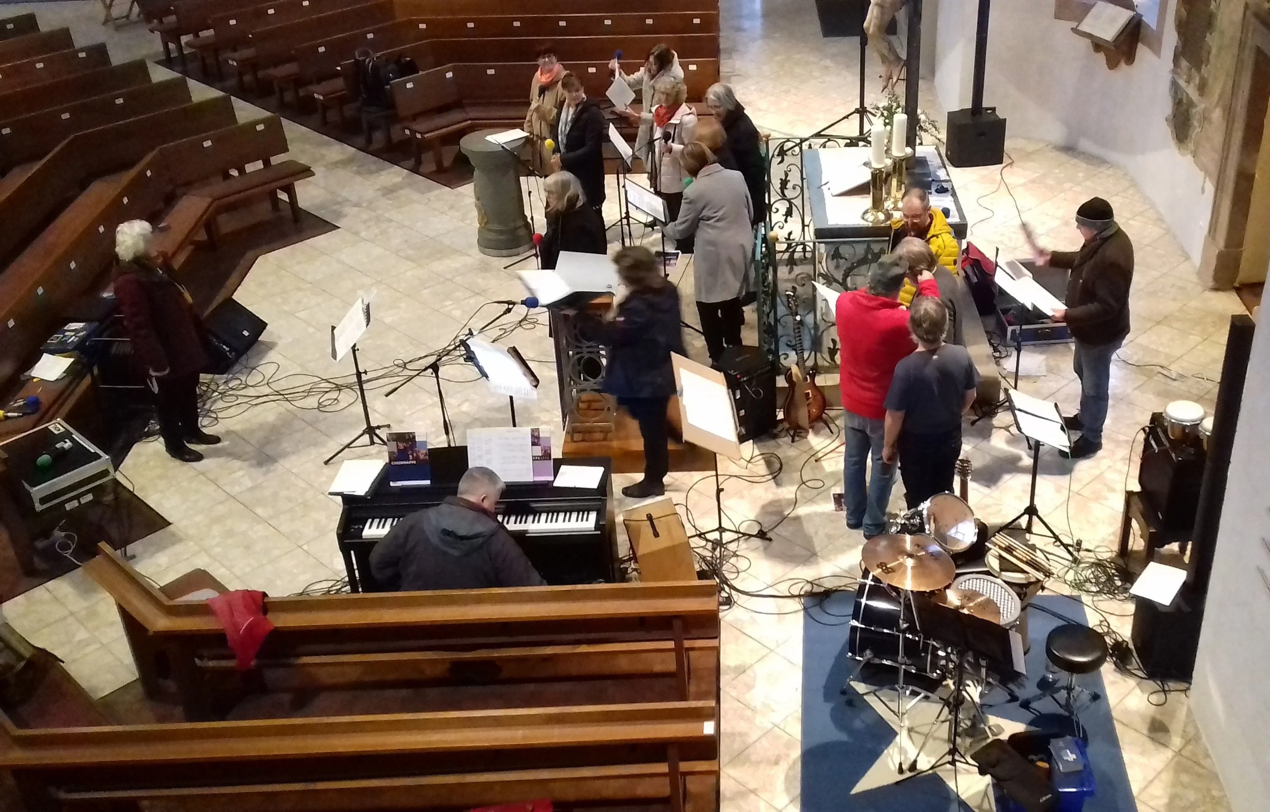 Mehrere Musiker spielen in einer Kirche, mit Klavier, Schlagzeug, Mikrofone und Notenständer, darunter weibliche und männliche Personen.
