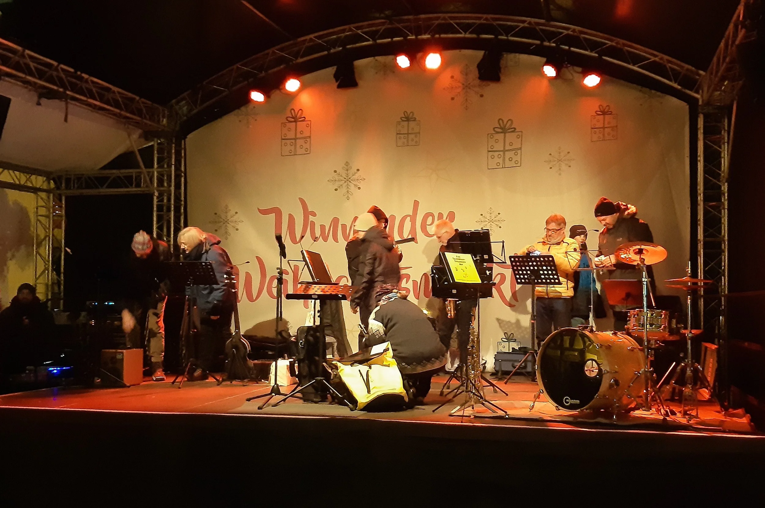 Musiker auf einer Bühne mit Weihnachtsdekoration im Hintergrund, rot beleuchtete Scheinwerfer, Instrumente und Musikzubehör sichtbar.