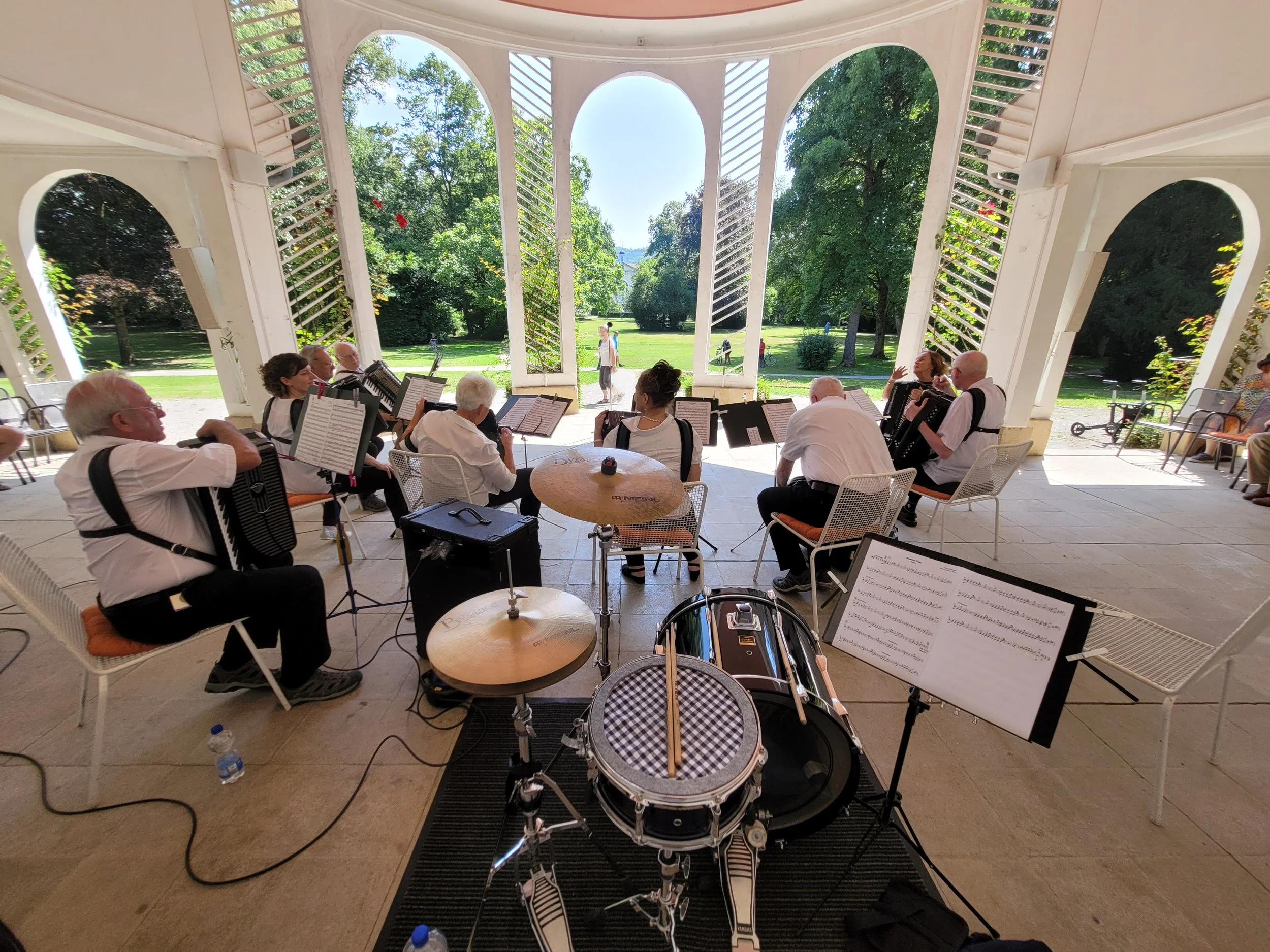 Eine Gruppe von Musikern spielt in einem offenen Pavillon im Park, mit Bäumen und Wegen im Hintergrund.