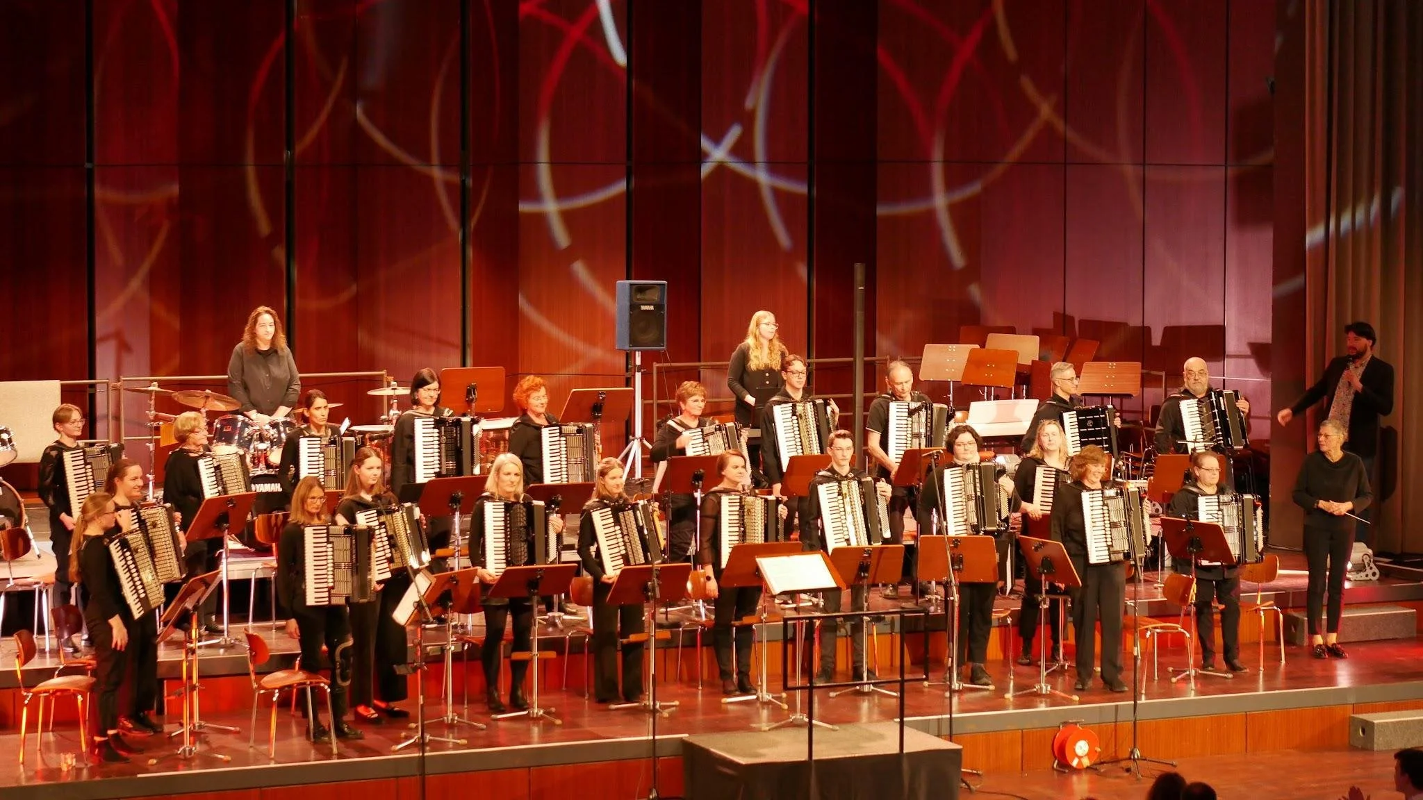 Ein Orchester auf einer Bühne mit Musikern, die Akkordeons spielen, und einem Dirigenten. Im Hintergrund eine Wand mit abstrakten Lichtern.