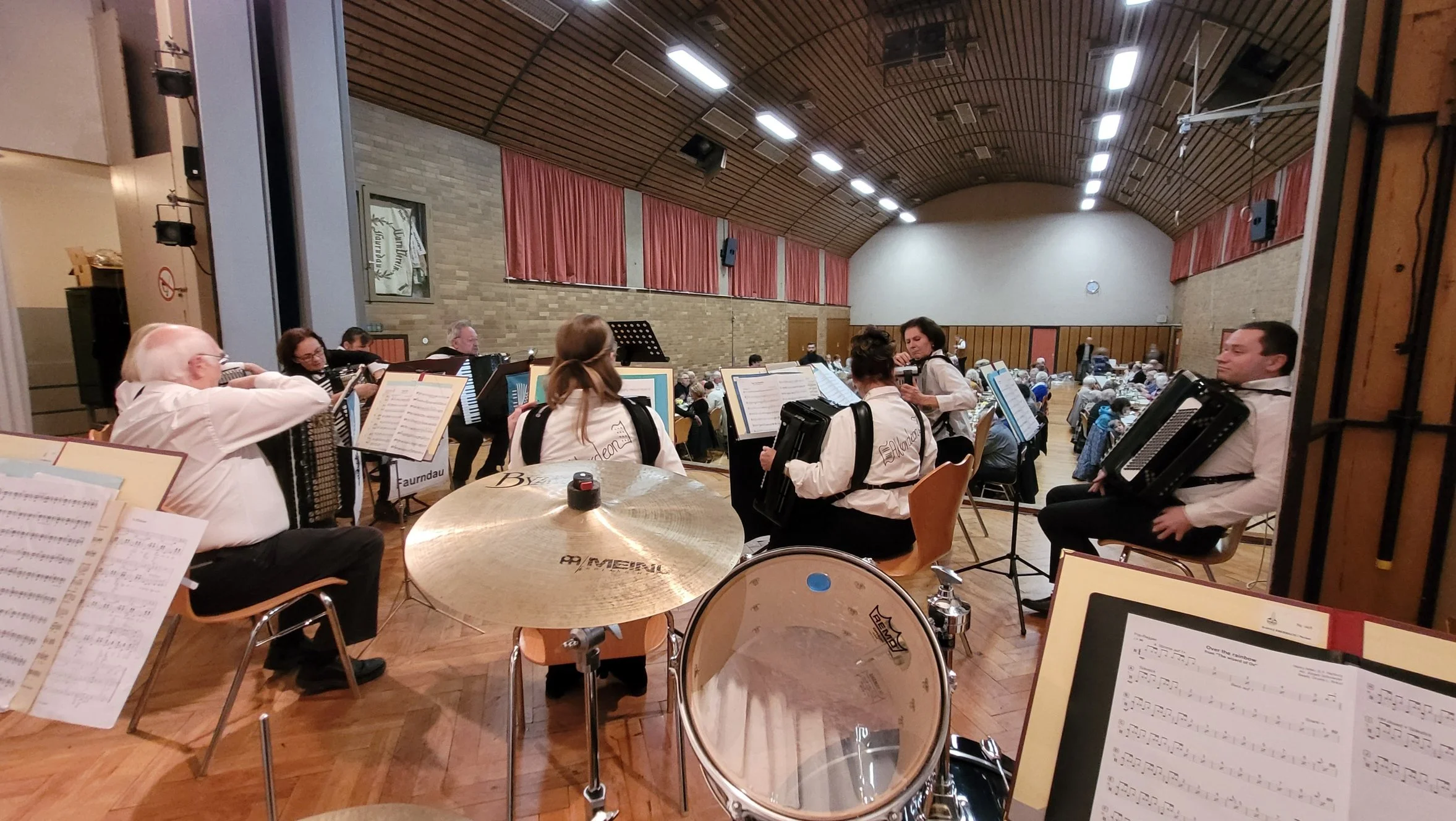 Ein Orchester spielt in einem Saal, mit Musikern, die Akkordeons, ein Schlagzeug, und andere Instrumente spielen, während das Publikum zusieht.