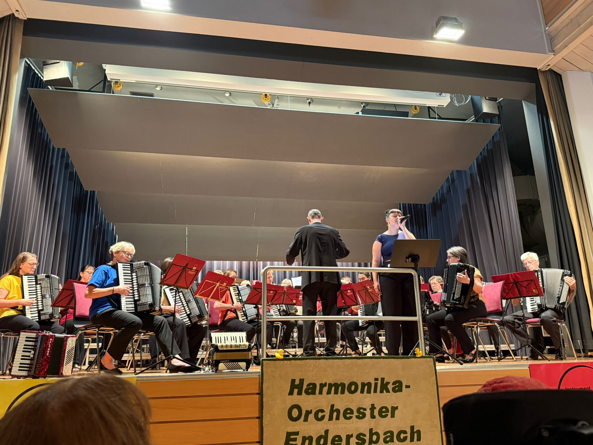 Ein Orchester auf einer Bühne während einer Aufführung, mit Musikern, die Instrumente wie Akkordeons spielen, und einem Dirigenten. Im Vordergrund ist ein Schild mit der Aufschrift 'Harmonik-Akkordeon-Orchester Endersbach' zu sehen.