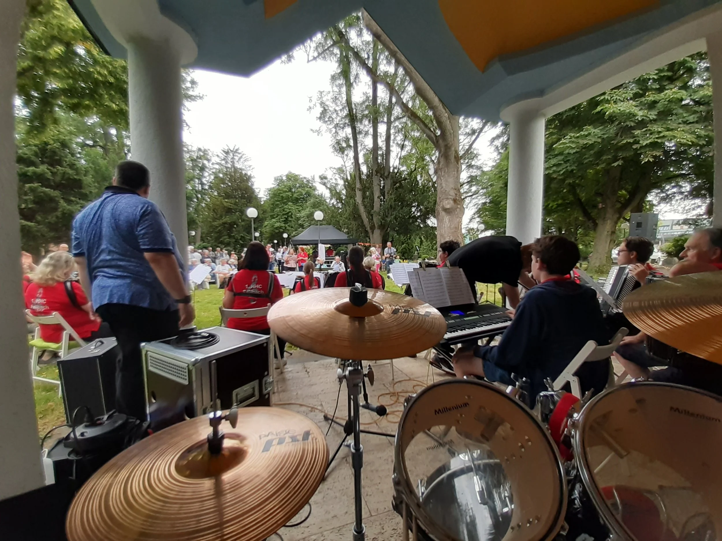 Musiker bei einem Open-Air-Konzert im Park, aufgenommen aus der Perspektive eines Schlagzeugers, mit mehreren Musiker auf der Bühne, viele Bäume im Hintergrund.
