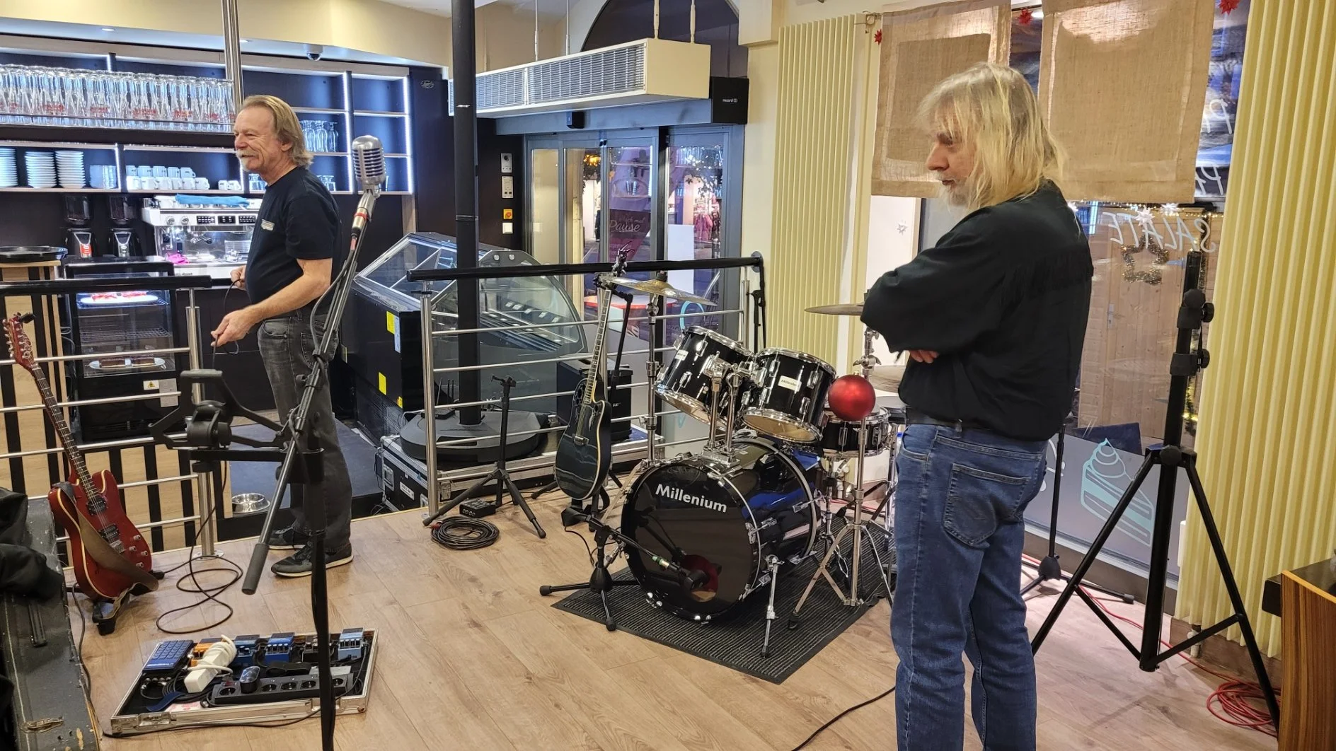Zwei Männer stehen in einem Musikraum mit Schlagzeug, Gitarren und anderen Musikinstrumenten. Einer der Männer trägt eine schwarze Jacke und Jeans, der andere einen schwarzen T-Shirt und graue Jeans. Es scheint, dass sie sich auf eine Musikperformance oder Probe vorbereiten.