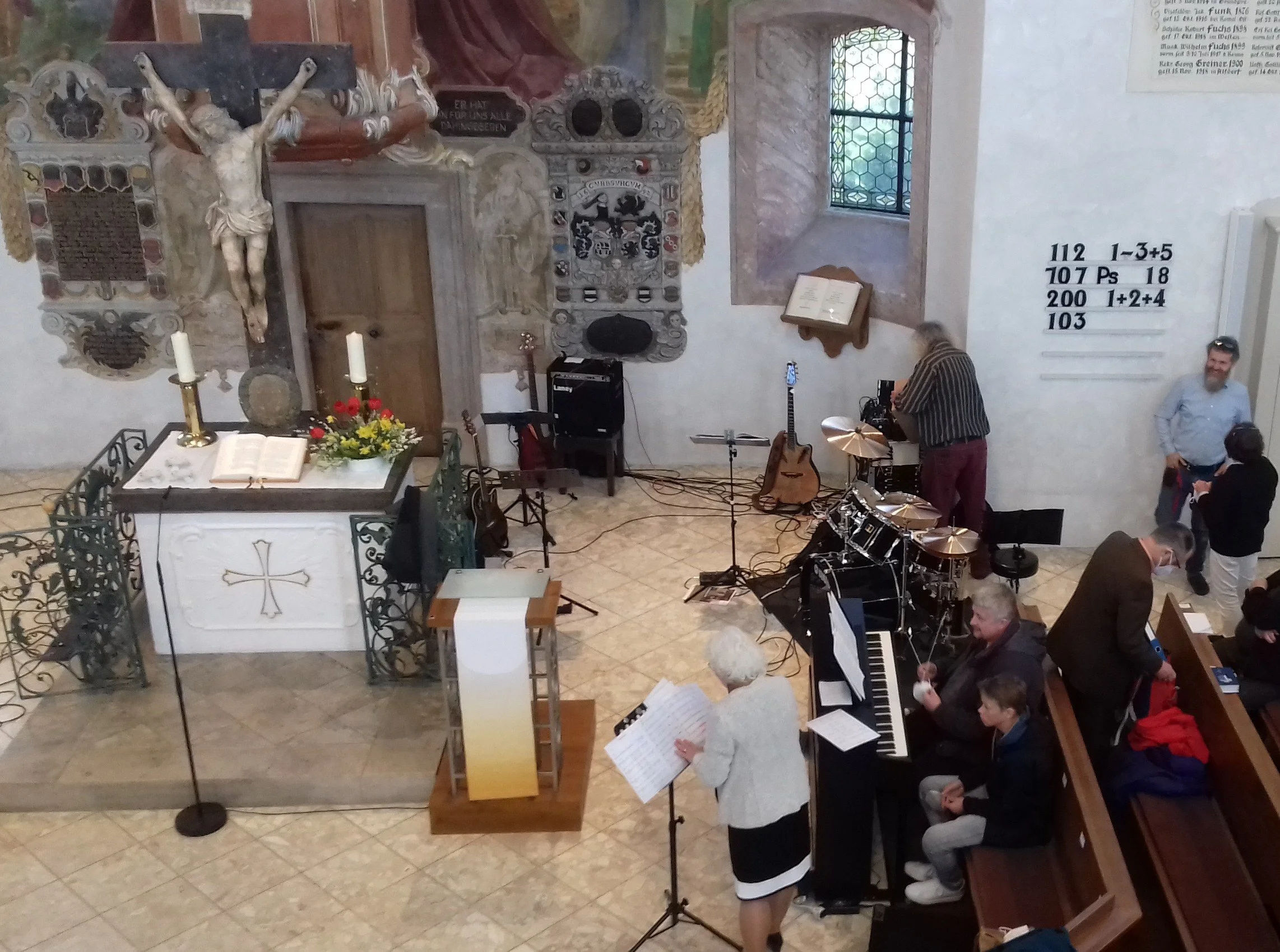 Das Bild zeigt eine Gruppe von Menschen, die in einer Kirche oder Kapelle Musik machen. Es gibt ein Podest mit einem aufgeschlagenen Liedbuch, Kerzen, Blumen und eine geschnitzte Figur Jesu am Kreuz. Die Musiker sind an Instrumenten wie Gitarre, Schlagzeug und einem Keyboard tätig. Es scheint eine Art Probe oder kleines Konzert zu sein.