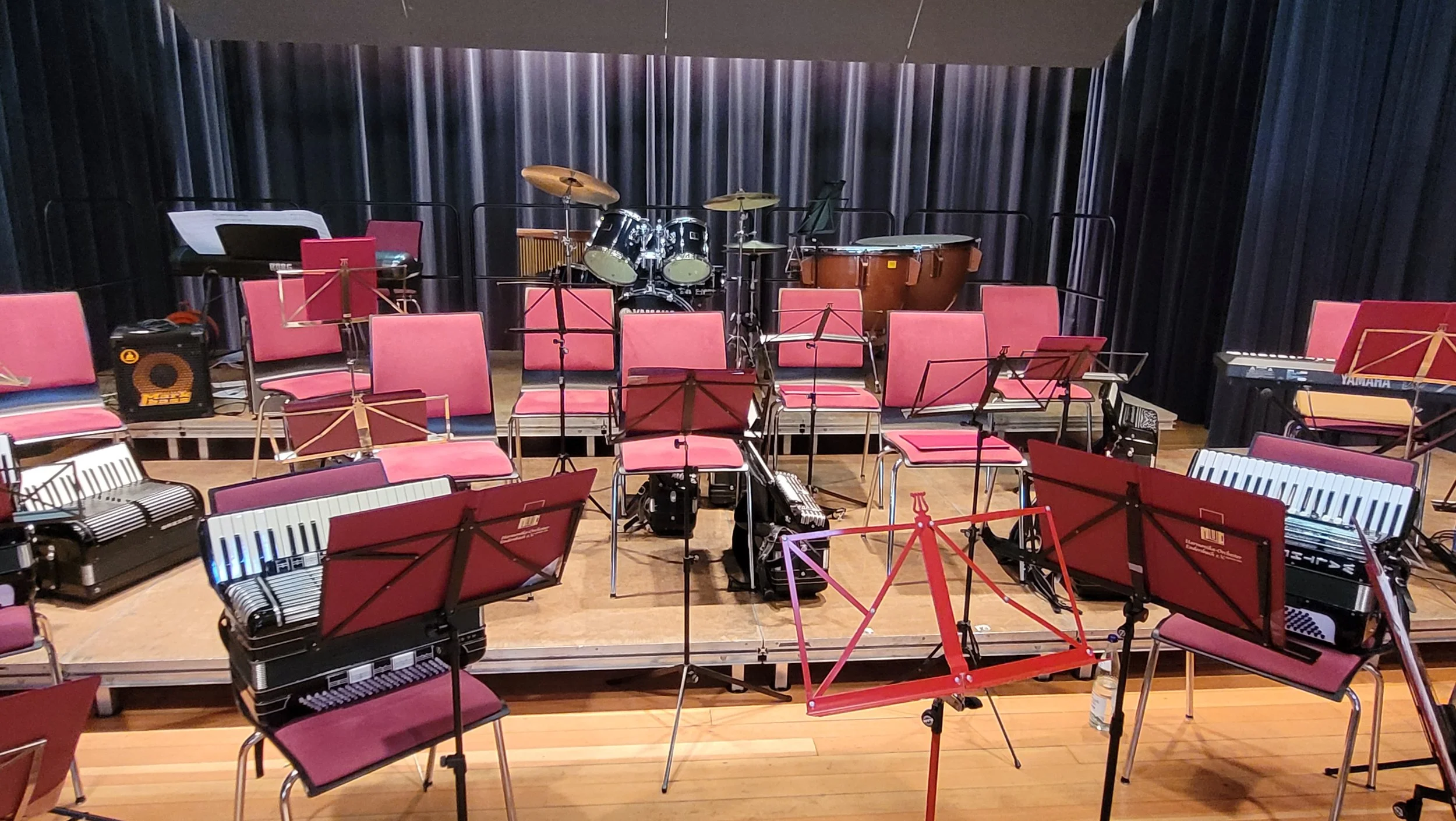 Leeres Orchester mit Musikinstrumenten, darunter Schlagzeug, Klaviere, Akkordeone und Notenständer, auf einer Bühne mit schwarzen Vorhängen im Hintergrund.