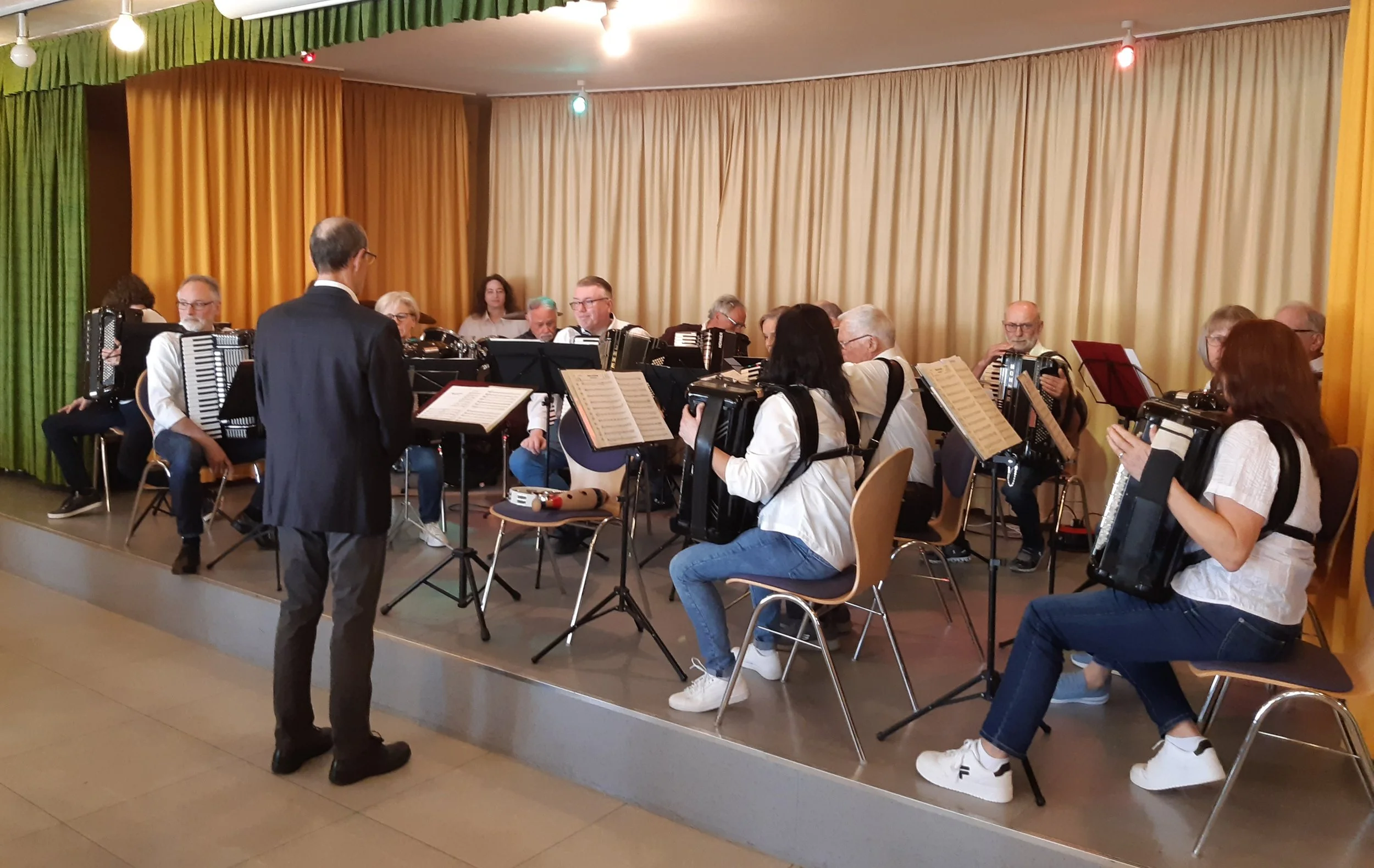 Eine Gruppe von Musikern spielt auf einer Bühne, dirigiert von einem Mann in einem schwarzen Anzug. Die Musiker sitzen mit Akkordeons, einige lesen Noten, im Hintergrund befinden sich goldene und grüne Vorhänge.
