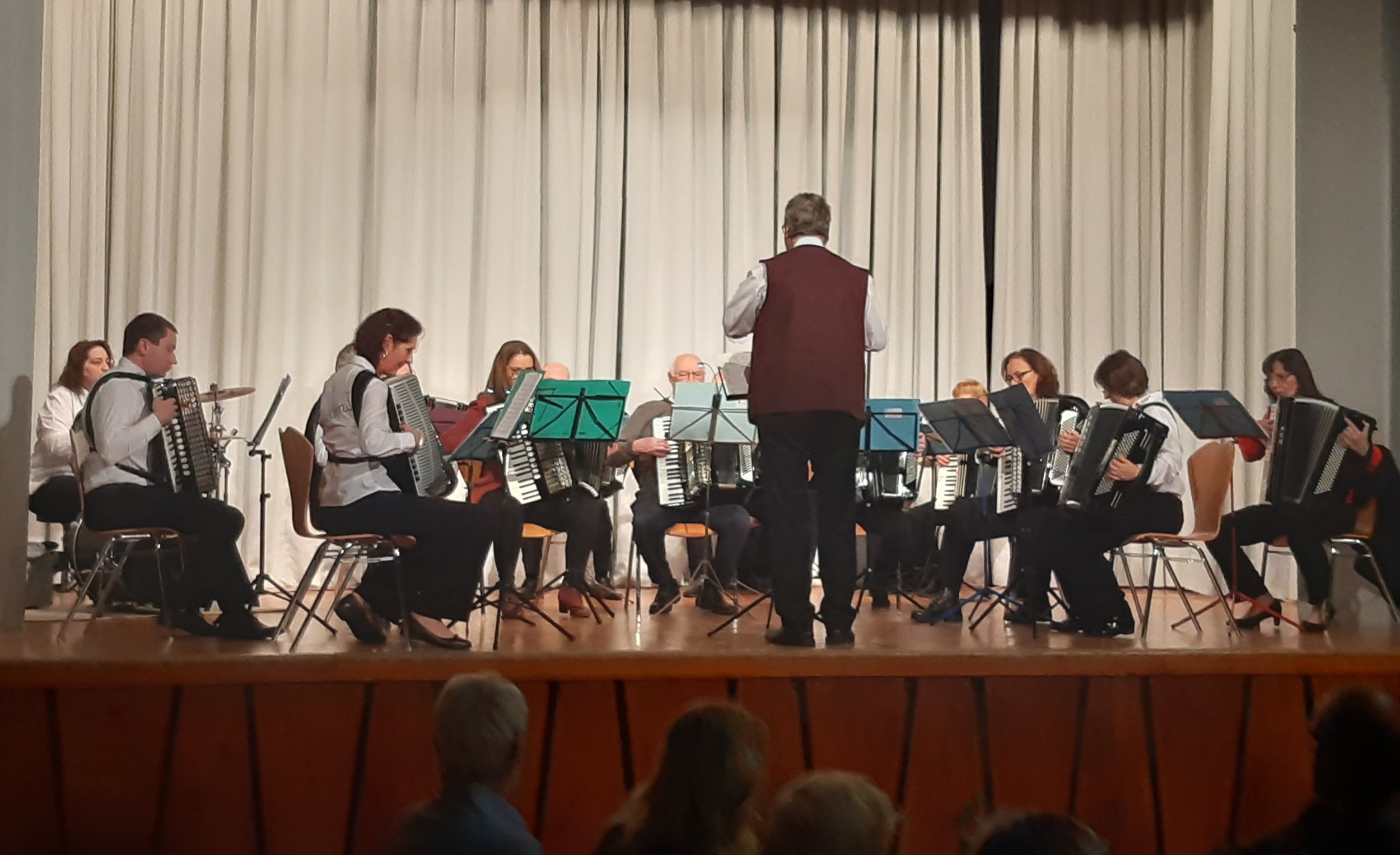 Ein Orchester auf einer Bühne mit einem Dirigenten in der Mitte. Die Musiker spielen Akkordeon und Schlagzeug.