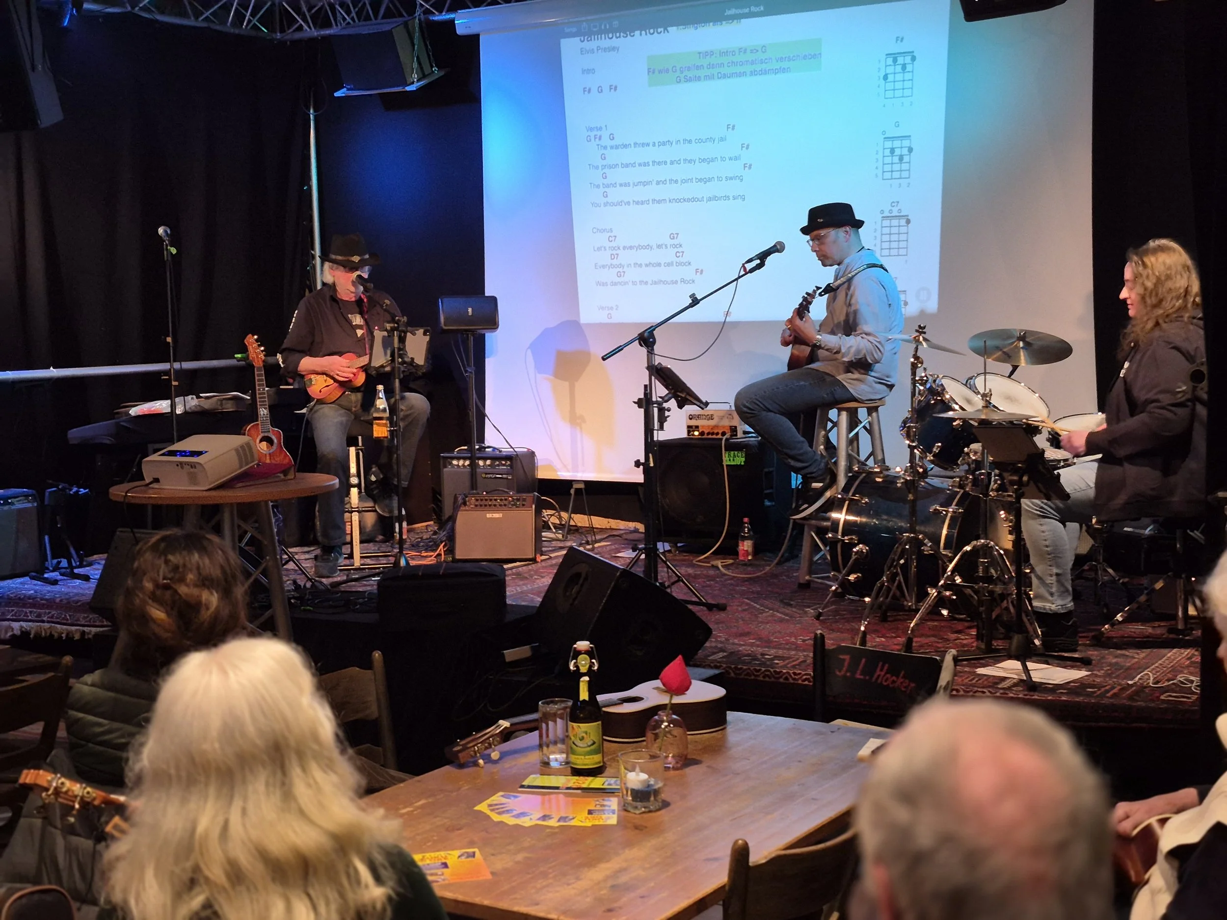 Musiker spielen auf einer Bühne während eines live-Events, im Hintergrund ein Bildschirm mit Liedtext und Gitarrengriffen, davor ein Publikum in einem dunklen Raum.