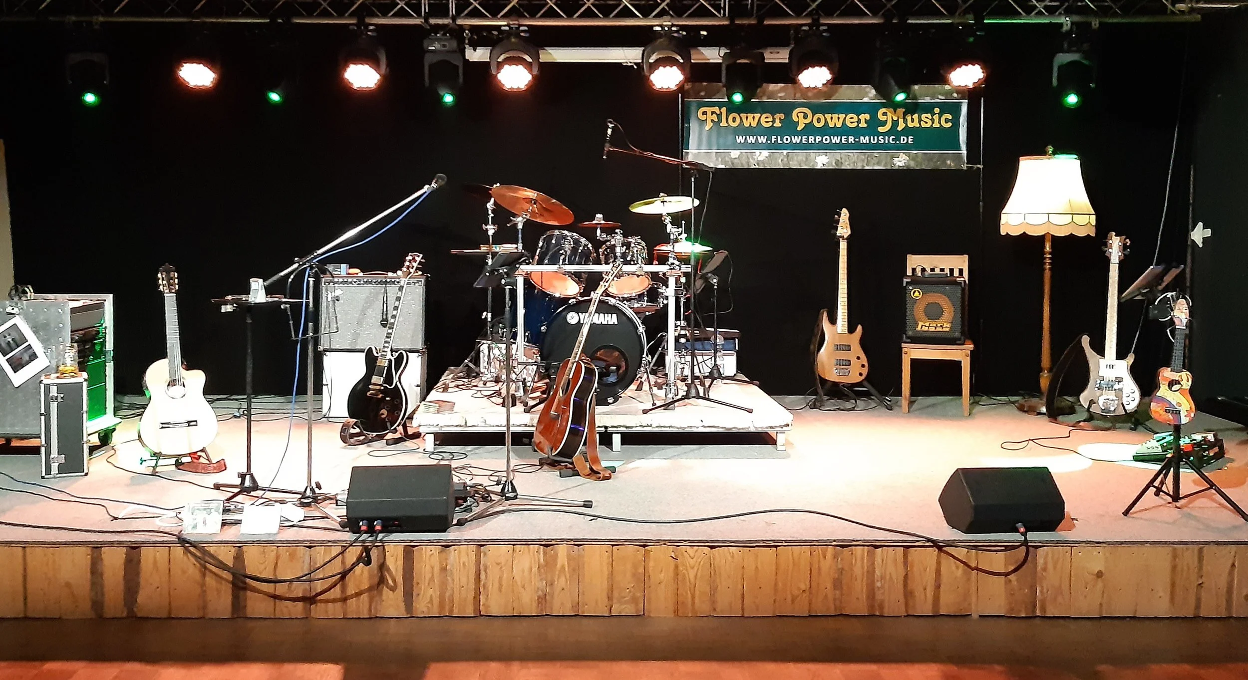 Eine leere Musikbühne mit verschiedenen Musikinstrumenten wie Gitarren, Schlagzeug und Verstärkern, dekoriert mit einem Lampenschirm und einem Banner mit der Aufschrift 'Flower Power Music'.