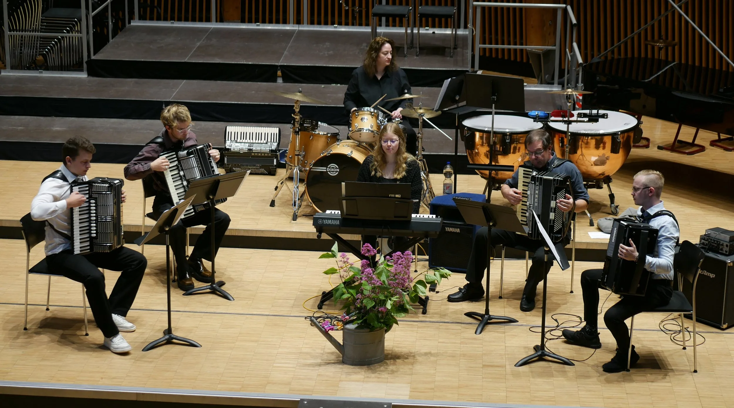 Eine Gruppe von sechs Musikern, die auf einer Bühne in einem Konzertsaal auftritt. Vier spielen Akkordeons, einer an einem Keyboard, eine Person an einem Schlagzeug.