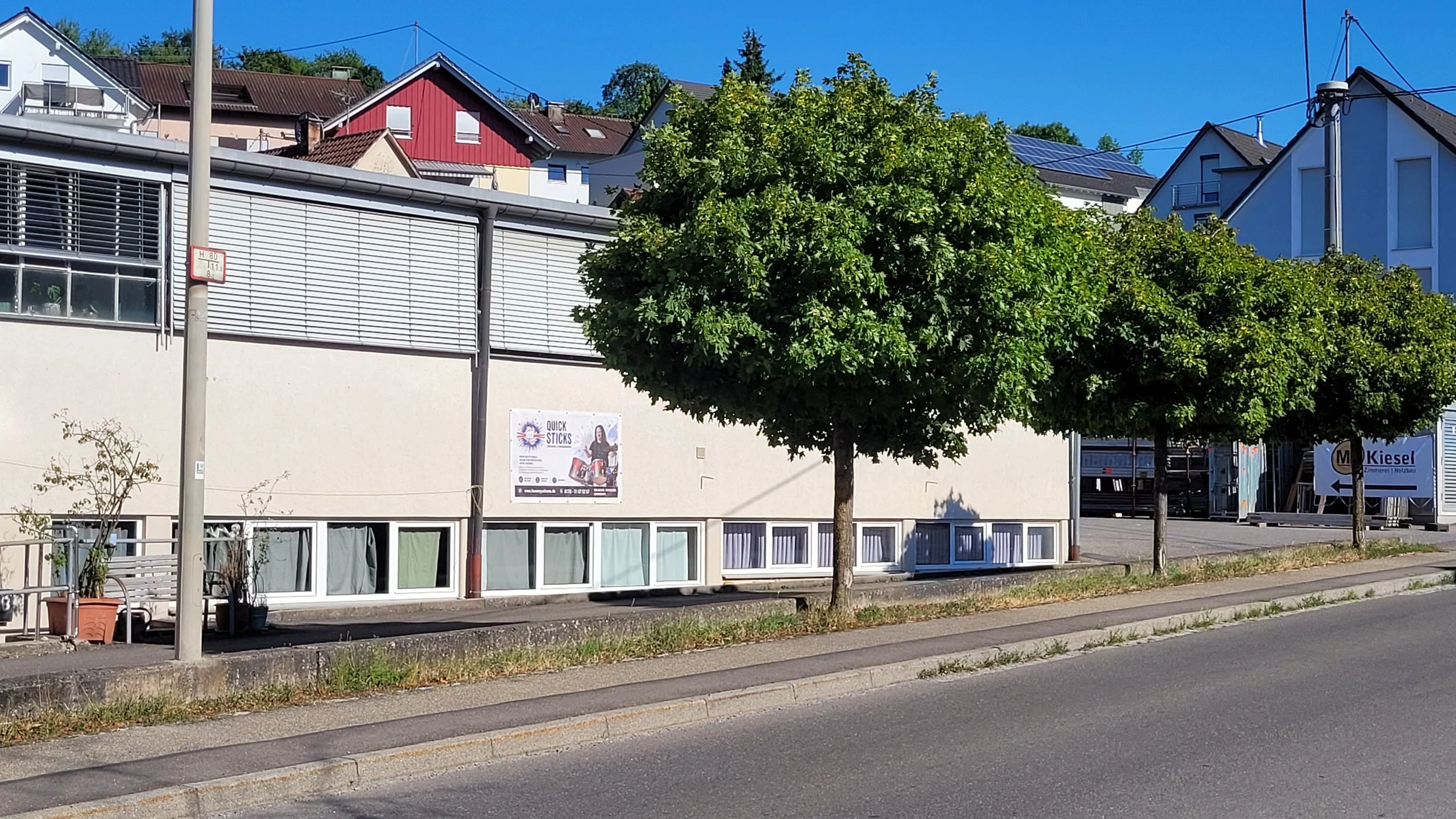 Straße mit Bäumen, Wohnhäuser und ein Geschäft mit einem Plakat, blauer Himmel.