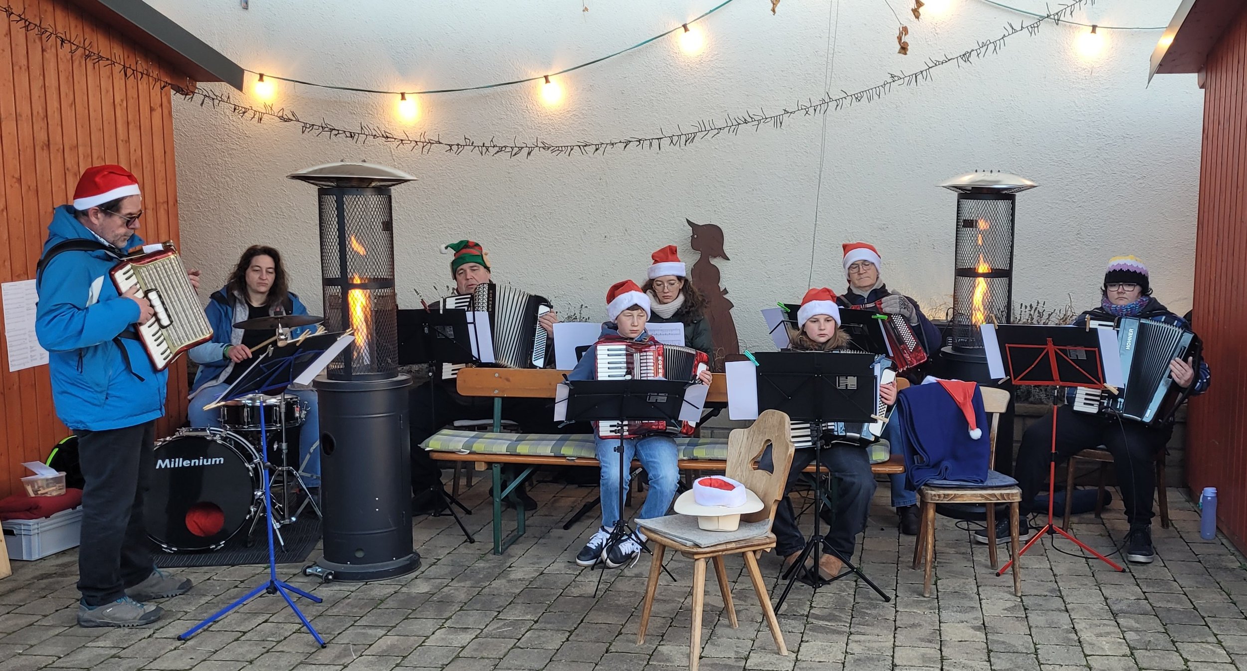 Eine Gruppe von Menschen, die Weihnachtslieder singt, spielt und zuhört. Sie tragen rote Weihnachtsmützen und einige haben kleine Elf-Hüte. Es gibt auch ein Kind. Das Bild wurde im Freien aufgenommen, mit Lichterketten an der Decke, Heizstrahlern und dekorativen Elementen im Hintergrund.